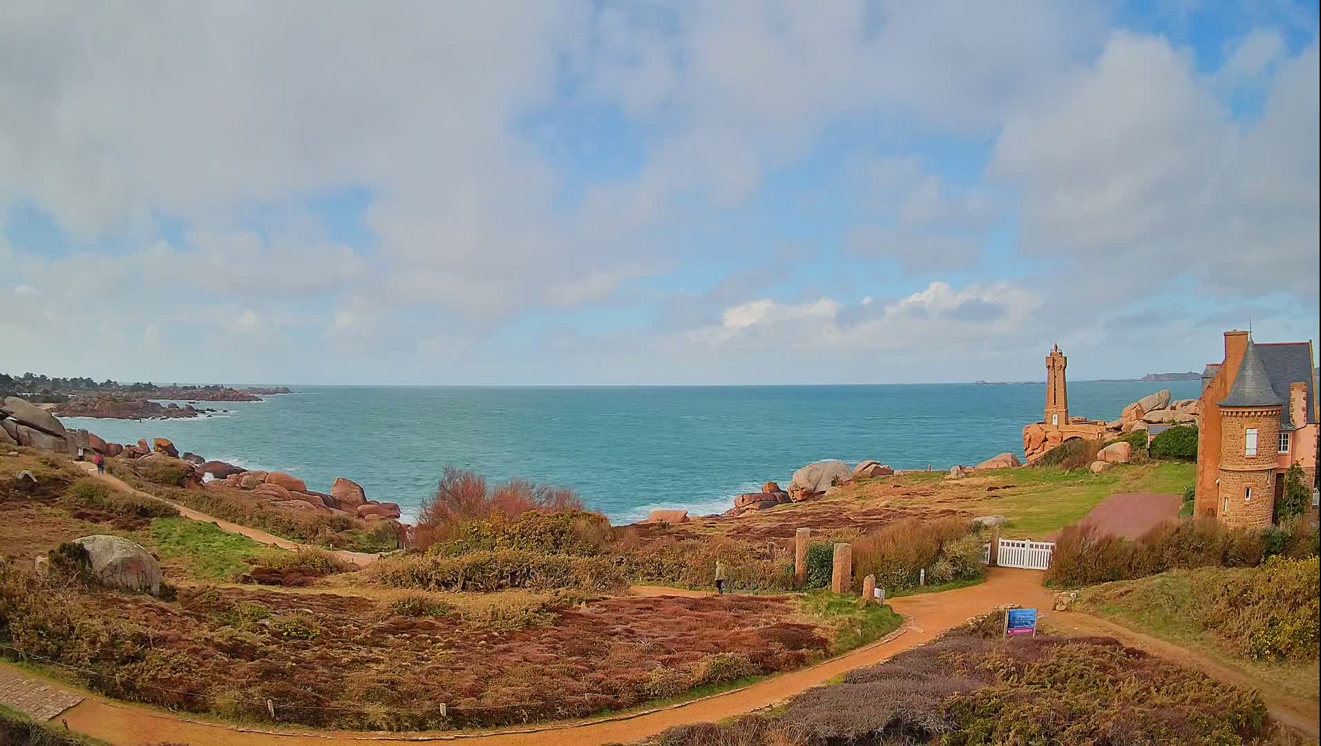 A coastal path winds through reddish-brown vegetation toward a stone tower and house overlooking a calm, blue ocean under a partly cloudy sky.
