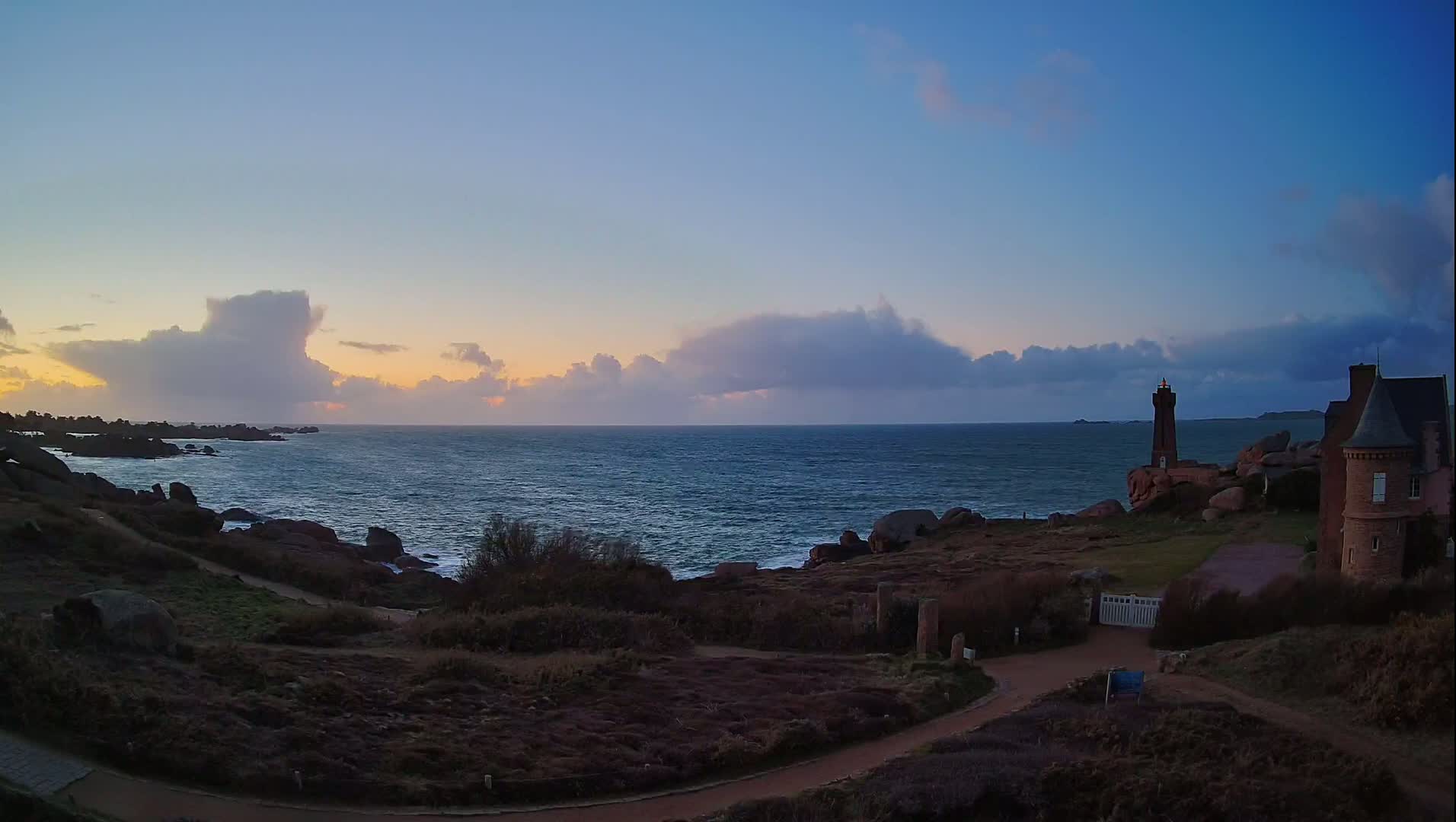 A rocky coastline with a lighthouse and stone building overlooks a choppy sea under a partly cloudy twilight sky.