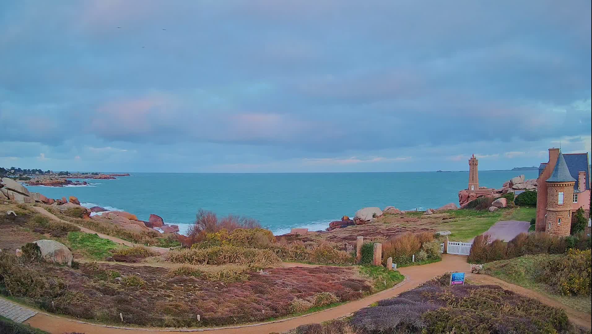 A coastal path winds through low-lying vegetation toward a stone building and lighthouse overlooking a calm, teal ocean under a mostly cloudy sky.