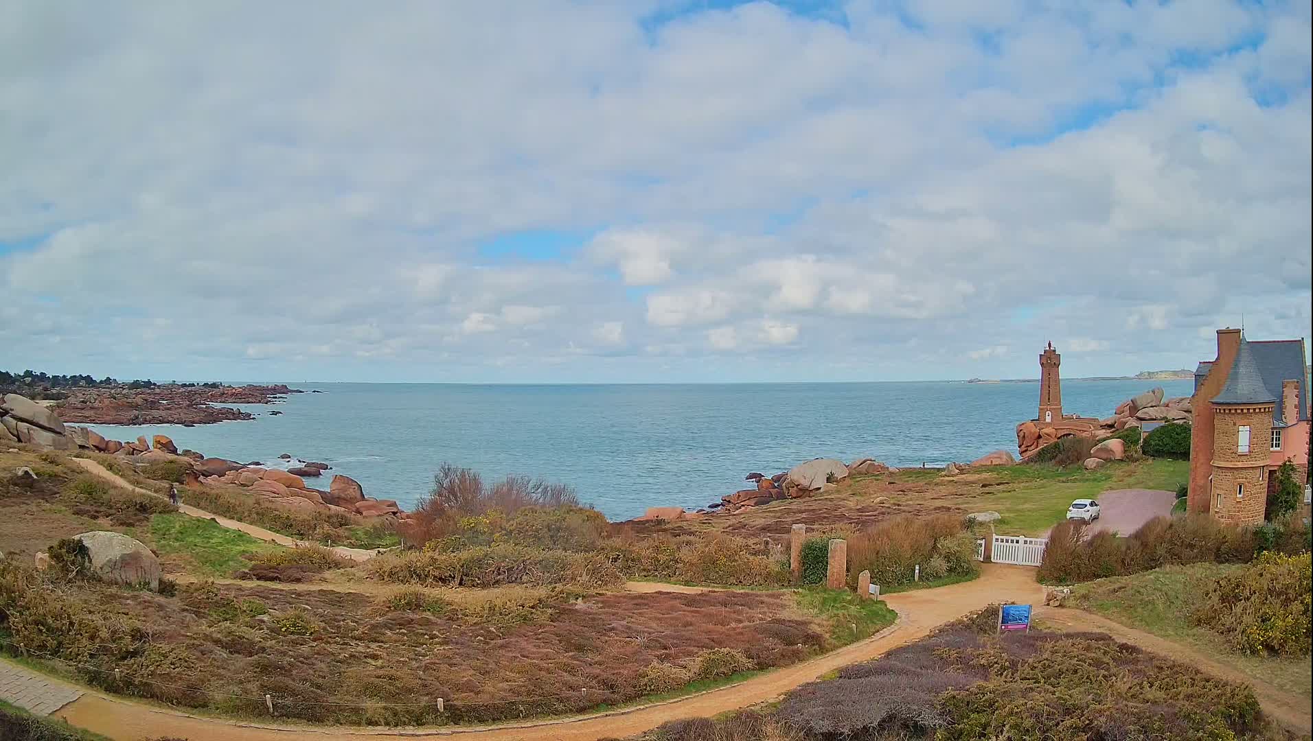 A coastal scene with a partly cloudy sky shows a lighthouse and a stone building overlooking a rocky shoreline and calm ocean.