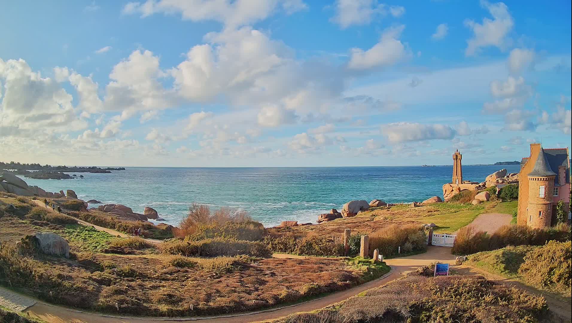A coastal scene under a partly cloudy blue sky shows a rocky shoreline, a lighthouse, a stone building, and winding paths, with people walking along the paths near the ocean.