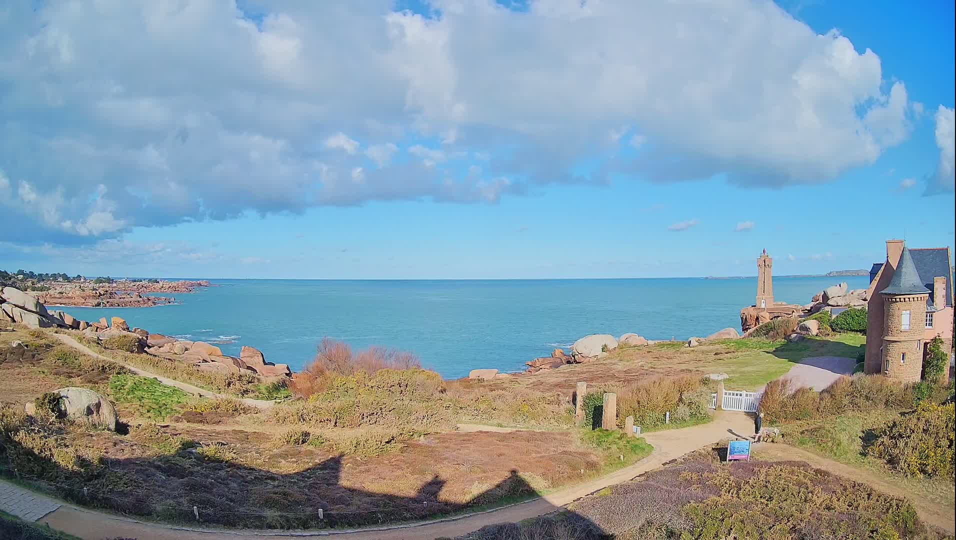 A partly cloudy blue sky overlooks a rocky coastline with a lighthouse and a stone house, alongside a dirt path.