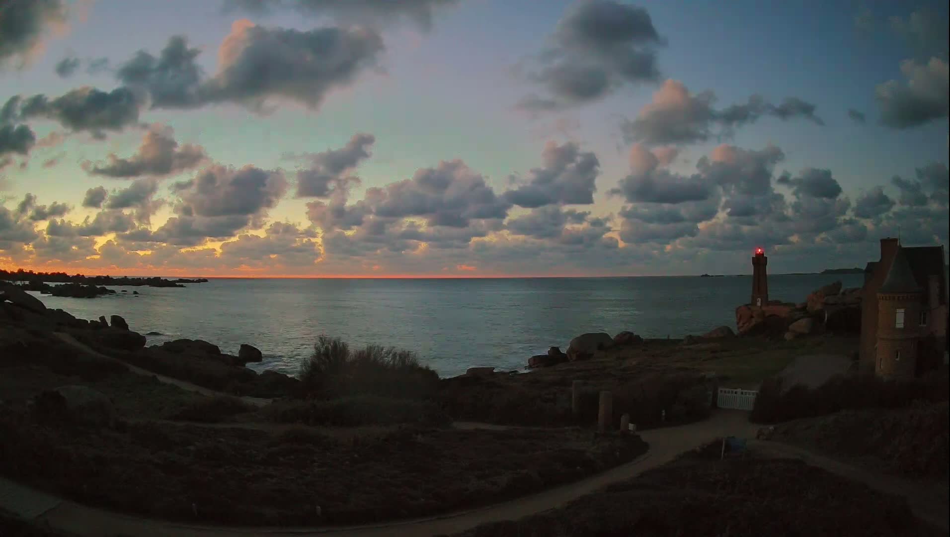 A rocky coastline at sunset under a cloudy sky features a lighthouse and a stone building.