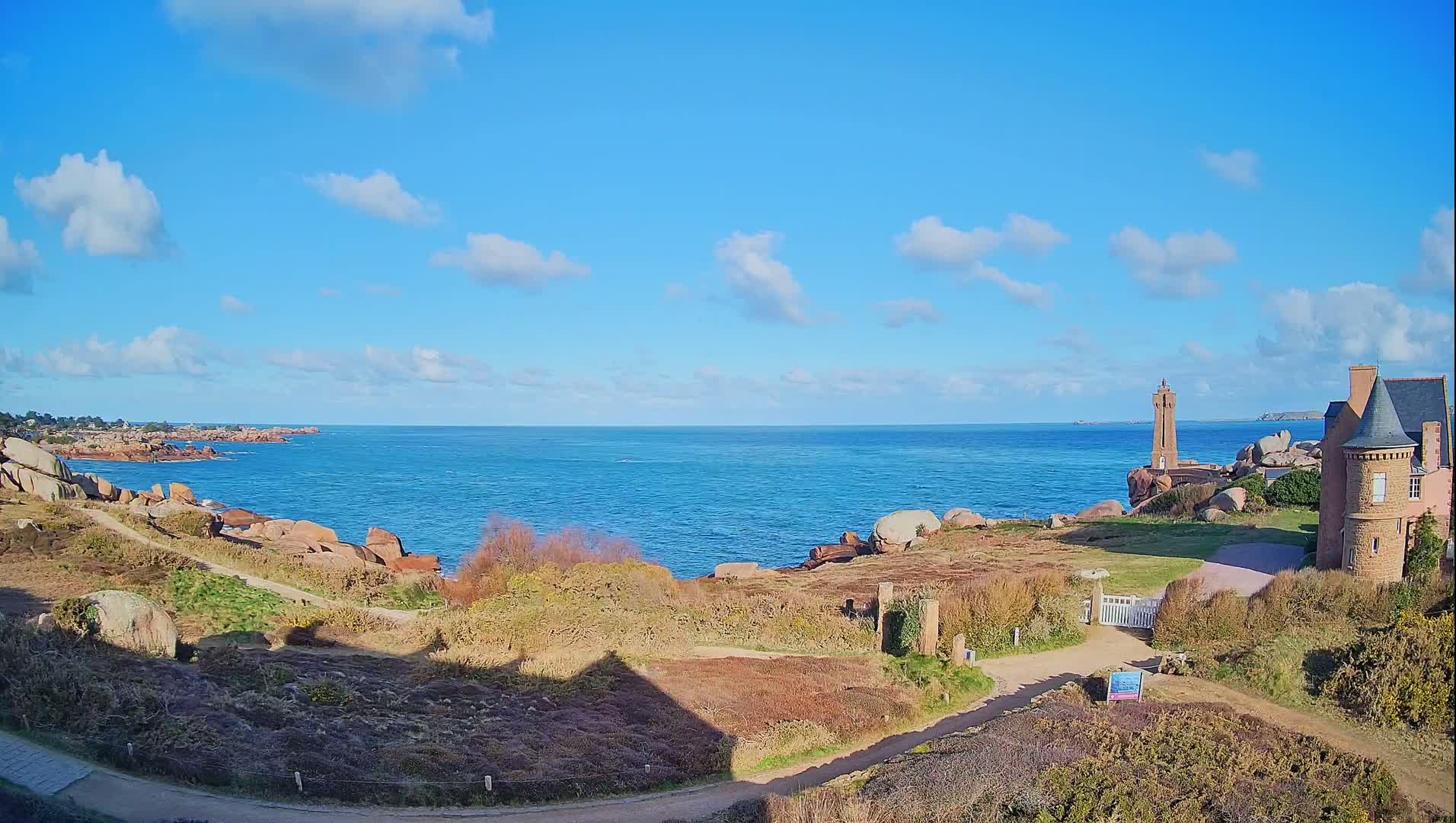 A coastal scene on a sunny day with a partly cloudy sky shows a stone lighthouse and a stone building near a rocky shoreline and blue ocean.