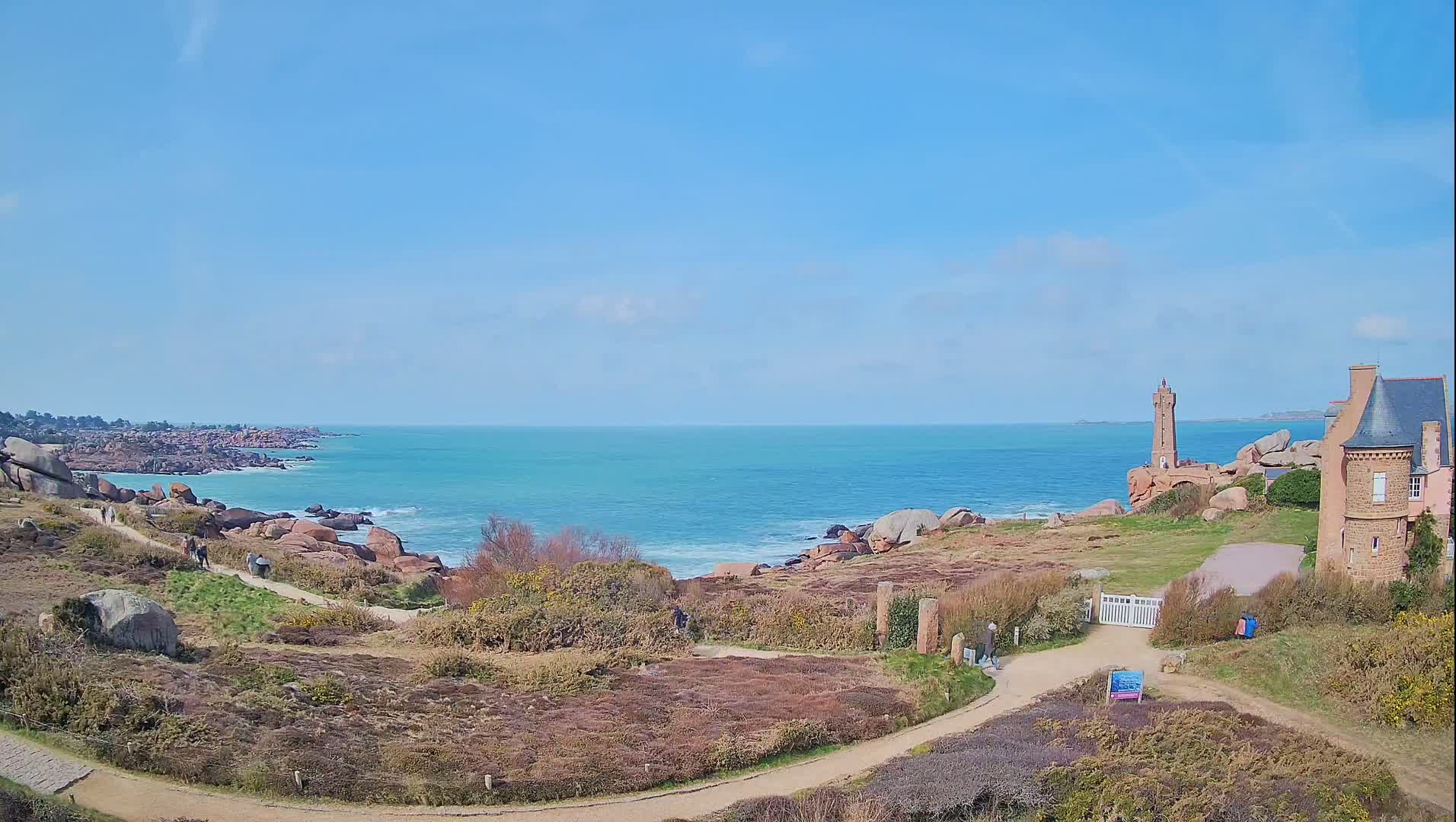 A coastal path winds through low-lying vegetation toward a lighthouse and house overlooking a calm, turquoise ocean under a clear blue sky.
