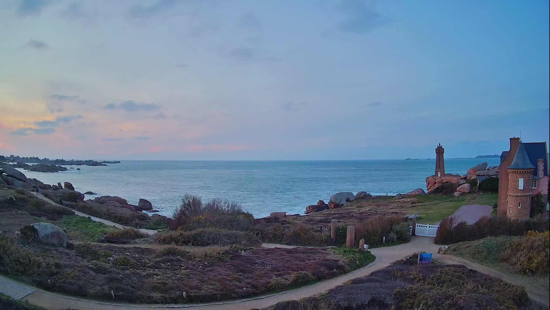 A coastal scene at sunset shows a calm sea, pink granite rocks, a lighthouse, a stone building, and winding paths amidst low-lying vegetation.