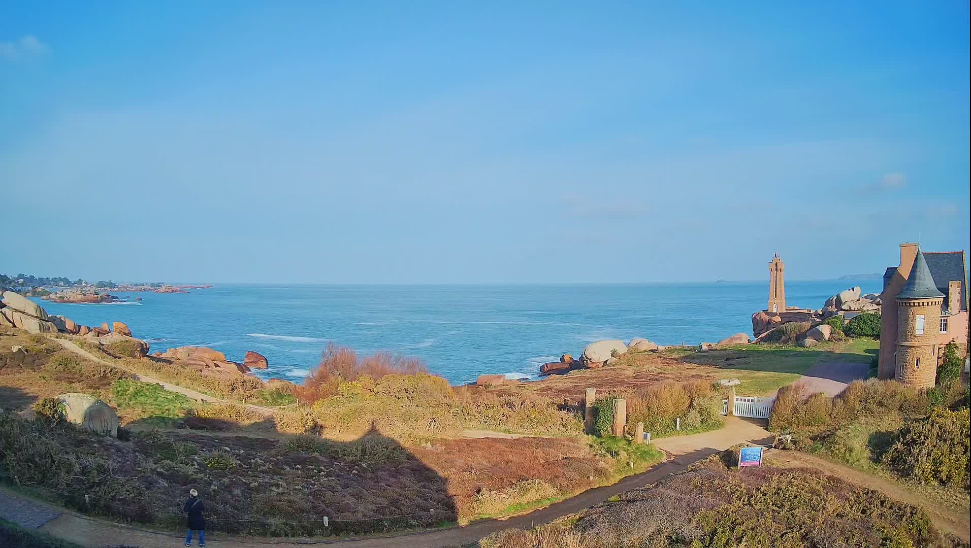 A person stands on a path overlooking a rocky coastline with a lighthouse and stone building under a clear blue sky.