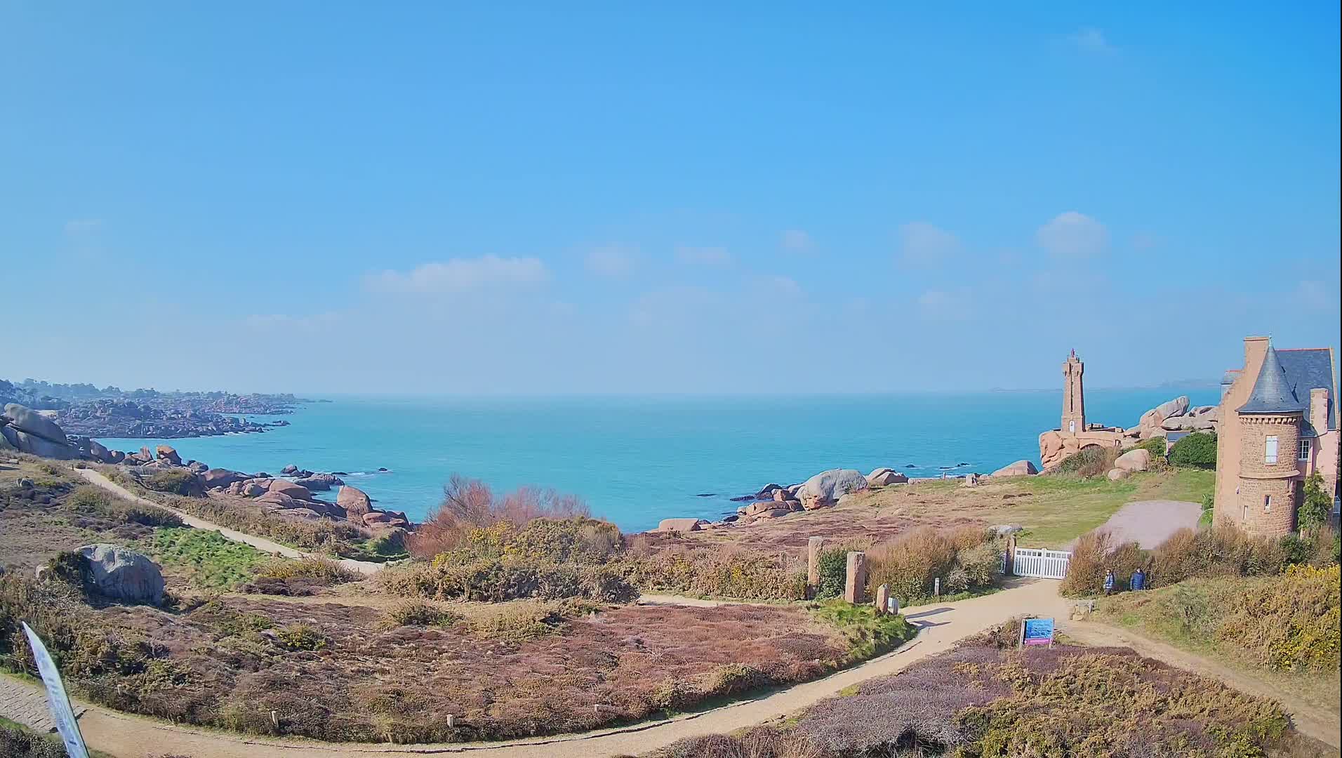 A coastal path winds through low-lying vegetation toward a stone building and a small lighthouse overlooking a calm, turquoise sea under a clear blue sky.