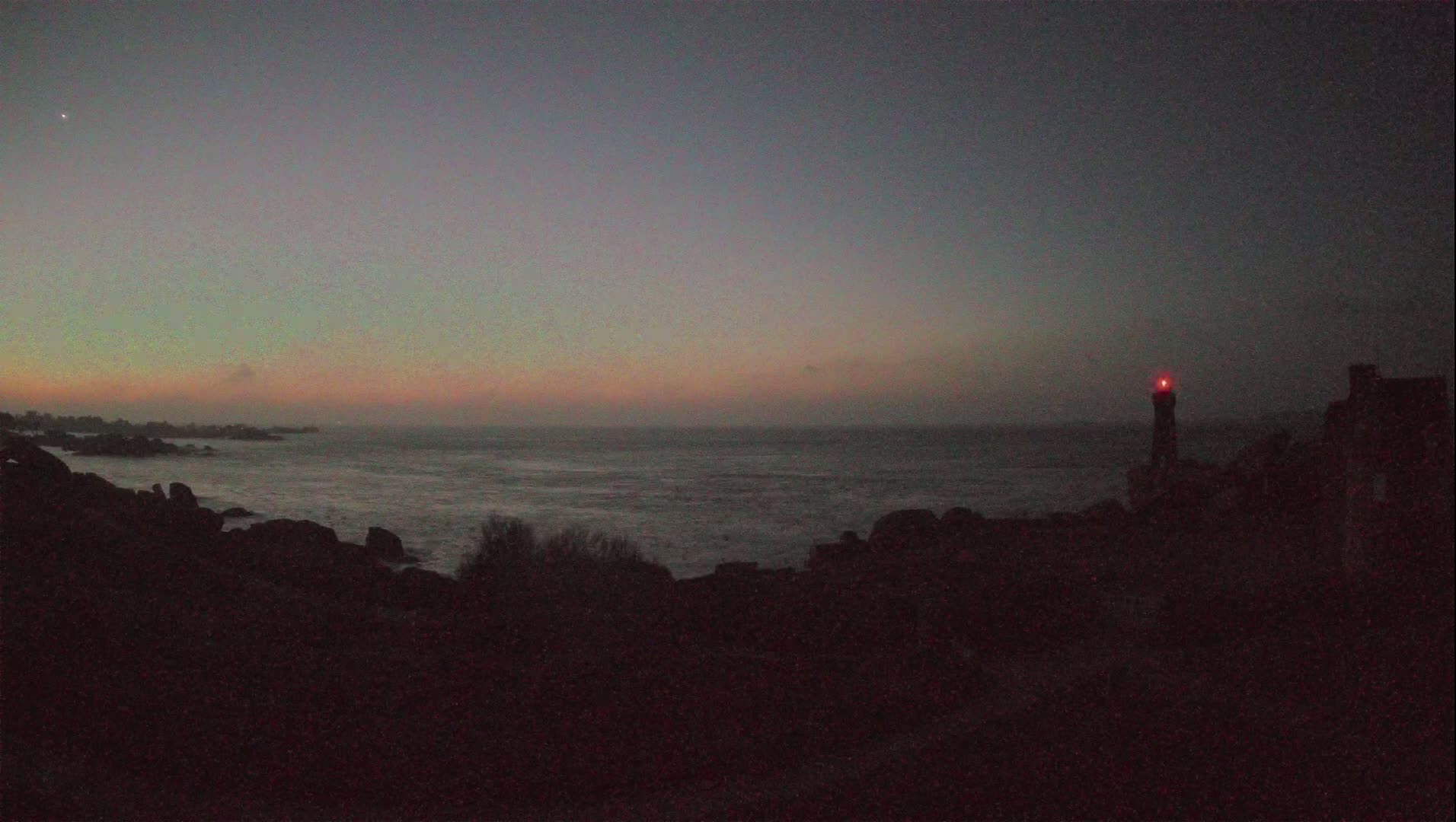 A rocky coastline at twilight, with a lighthouse emitting a red light in the distance, under a pale, pastel sky.