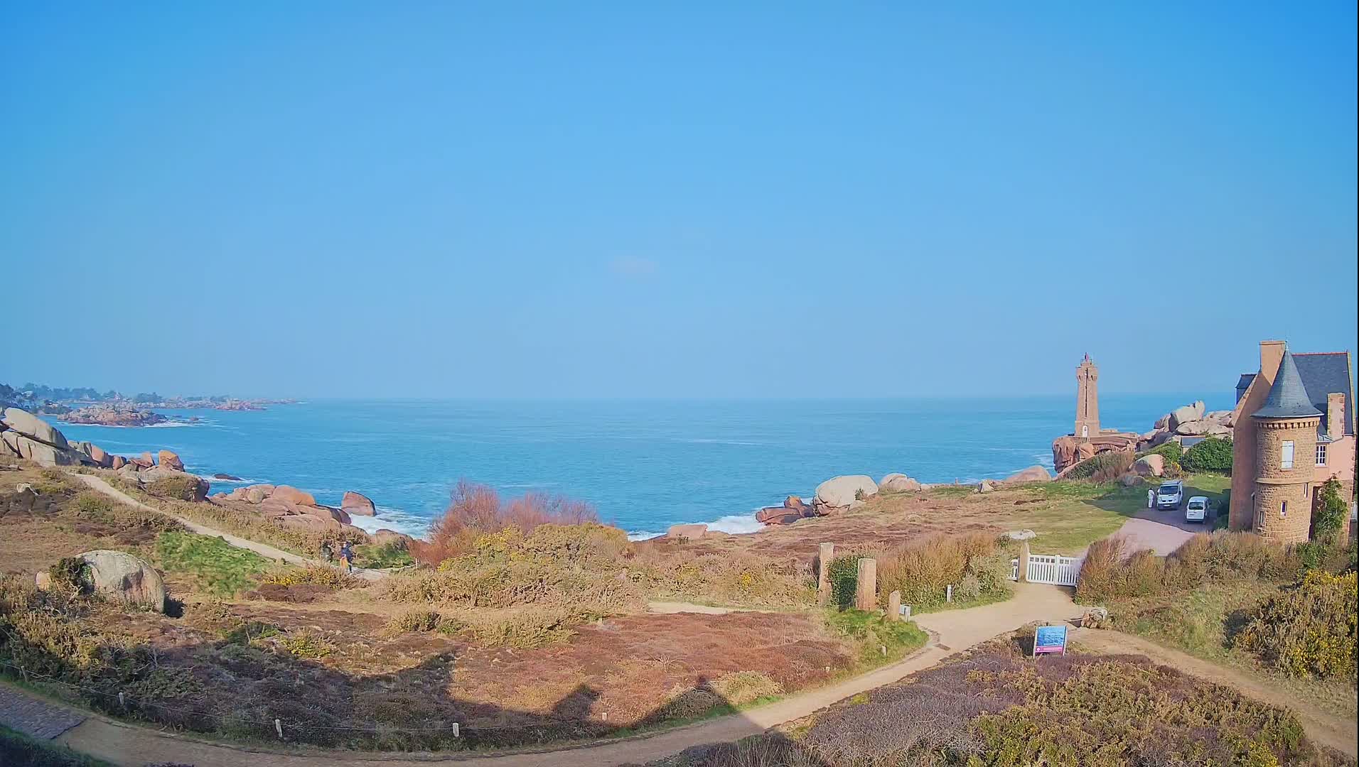A coastal scene on a clear, sunny day shows a rocky shoreline with a lighthouse and a stone building near a path leading to the ocean.