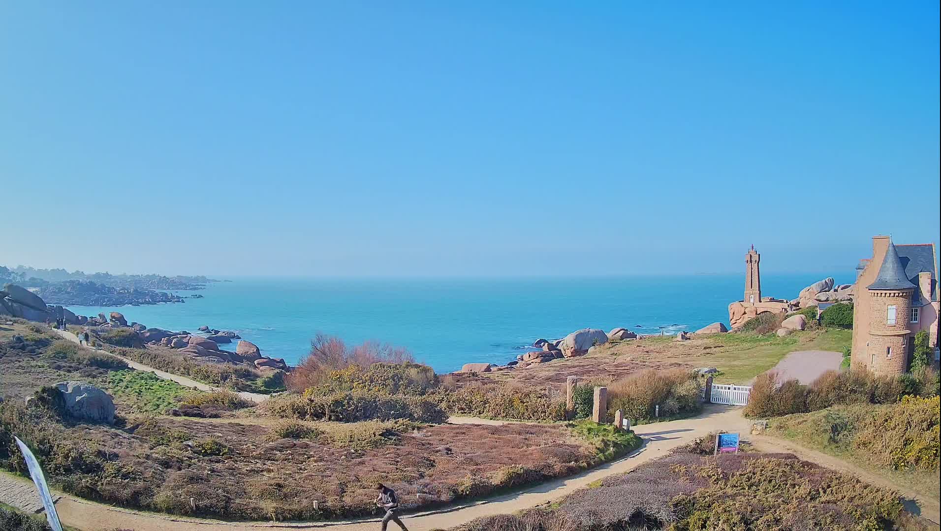 A person walks along a coastal path near a stone building and lighthouse overlooking a calm, turquoise ocean under a clear blue sky.