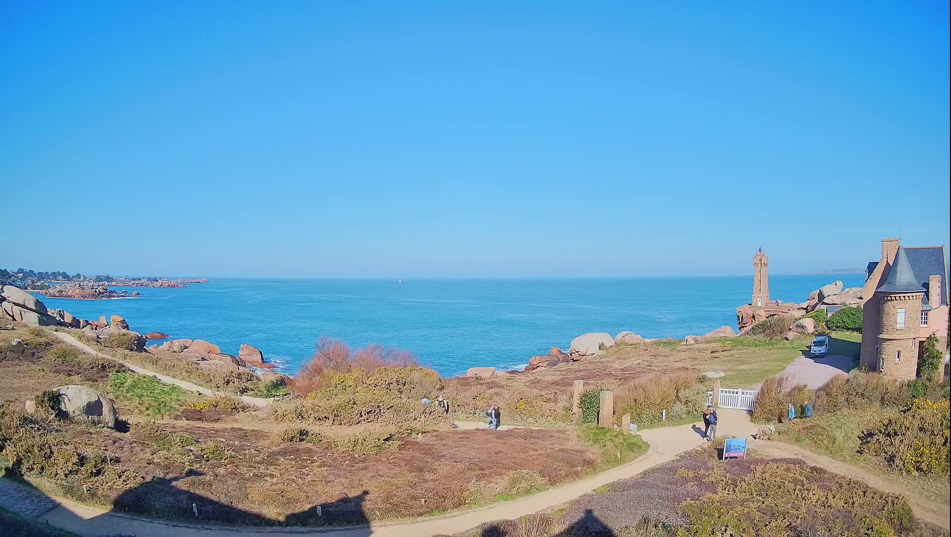 A coastal scene on a sunny day shows a stone lighthouse and building near a rocky shoreline and calm blue ocean.
