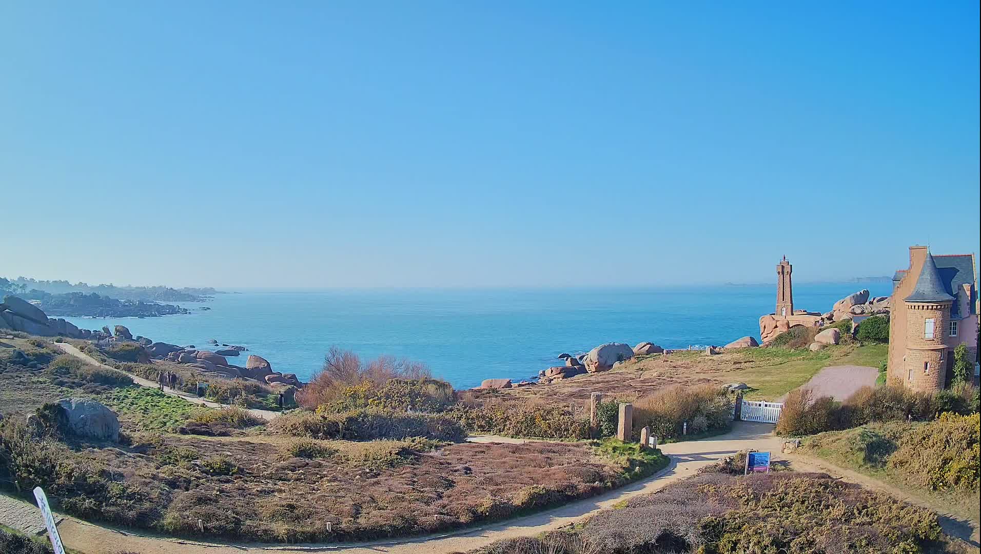 A coastal path winds through low-lying vegetation towards a stone building and a small lighthouse overlooking a calm, blue ocean under a clear, sunny sky.