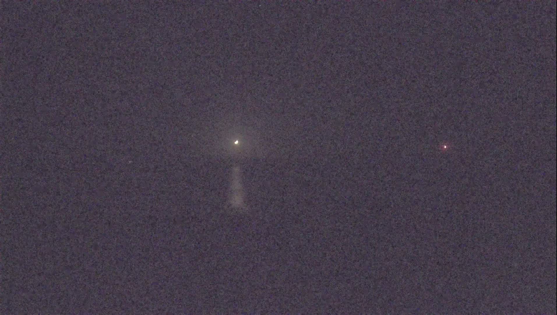 A bright light with a faint trail and a dim red light are visible in the night sky.