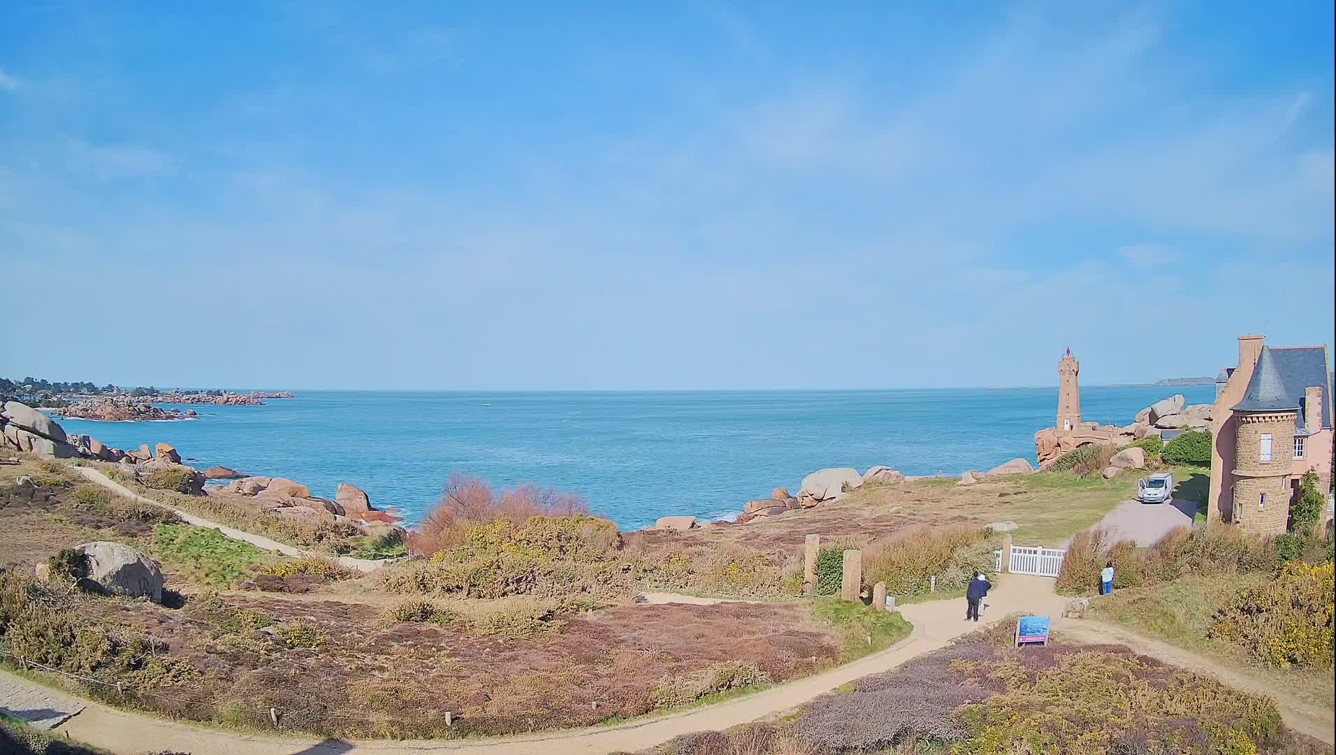 A coastal scene on a sunny day shows a lighthouse and a stone building overlooking a calm ocean, with a pathway winding through rocky terrain and sparse vegetation.