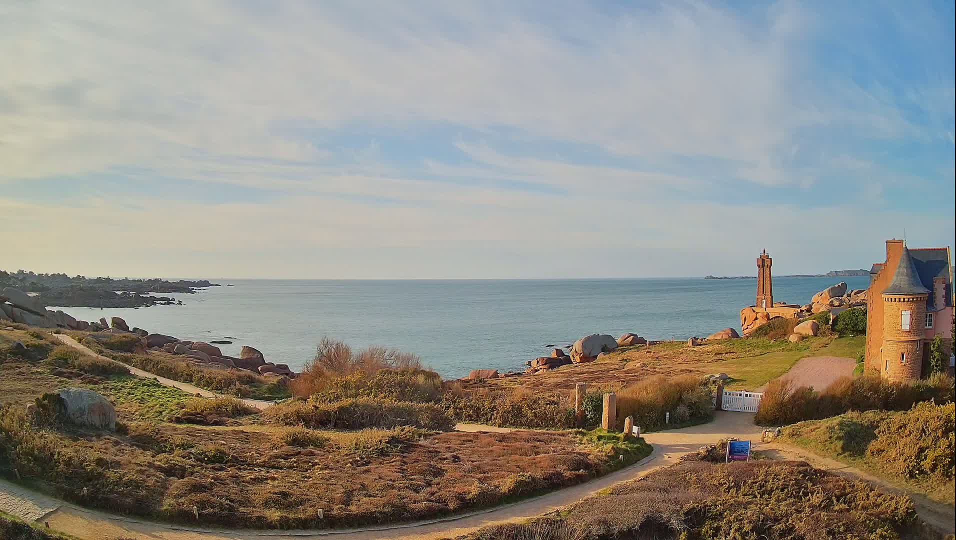 A coastal scene under a partly cloudy sky features a stone lighthouse and building overlooking a rocky shoreline and calm ocean.