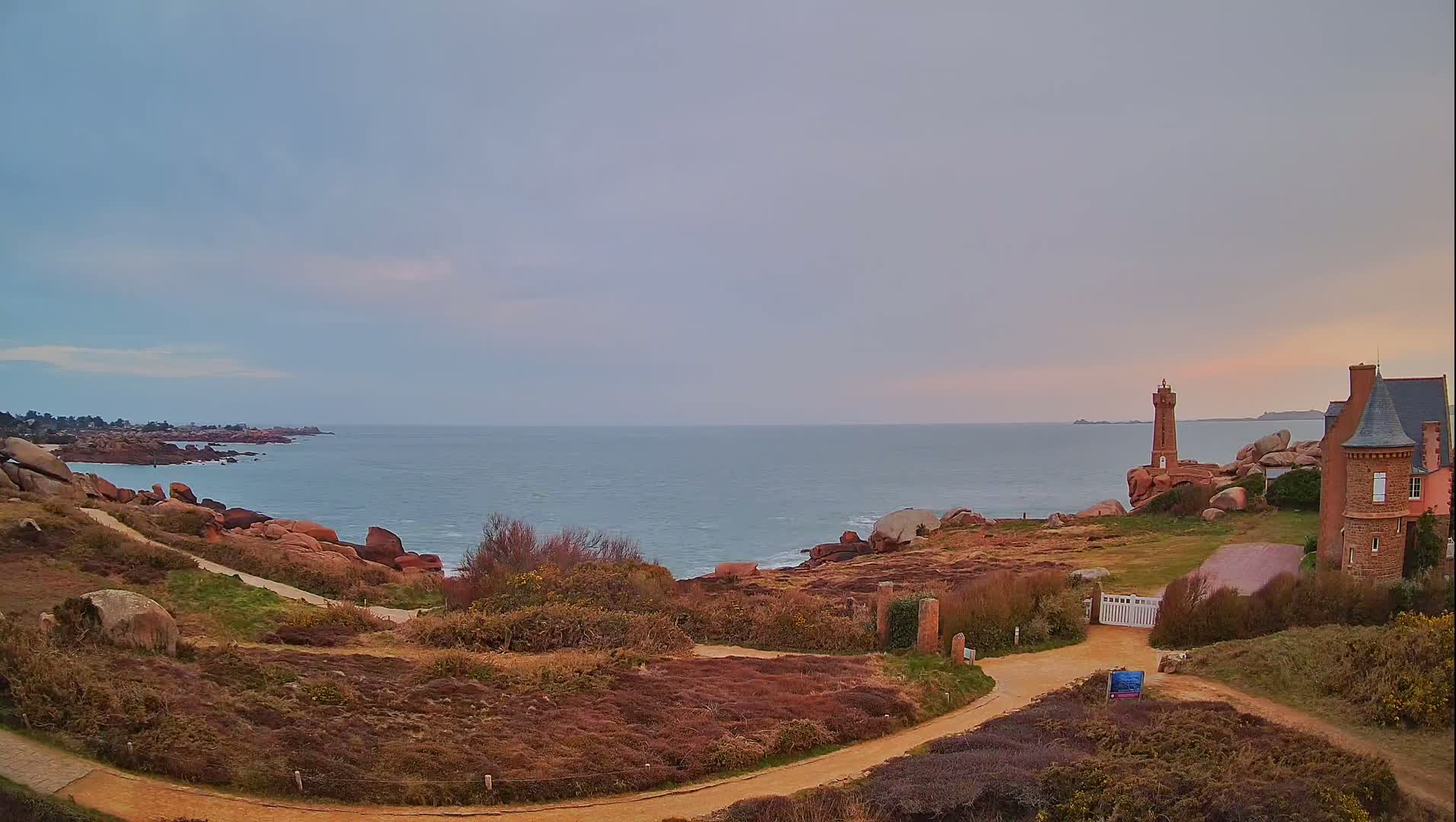A coastal path winds through low-lying vegetation towards a brick lighthouse and house overlooking a calm sea under a cloudy sky.