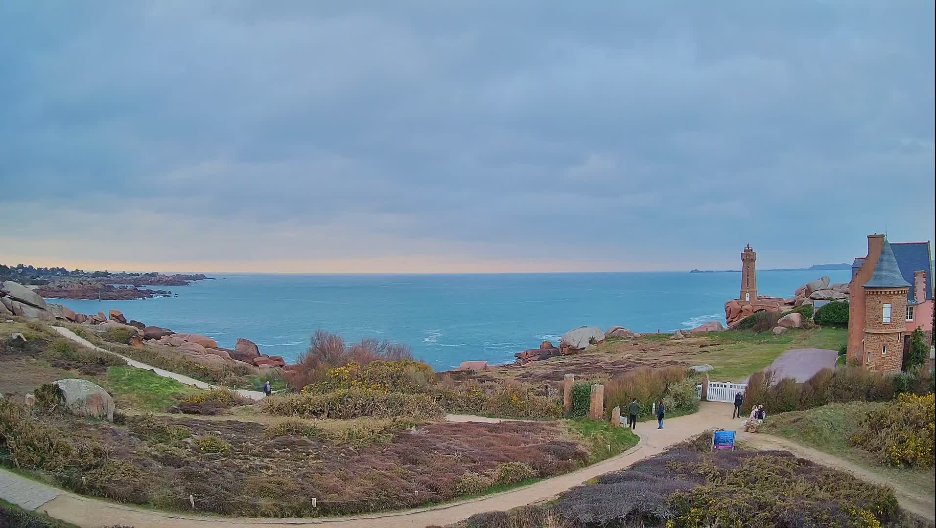 A coastal path winds through low-lying vegetation toward a lighthouse and a stone building overlooking a calm, blue ocean under a cloudy sky.