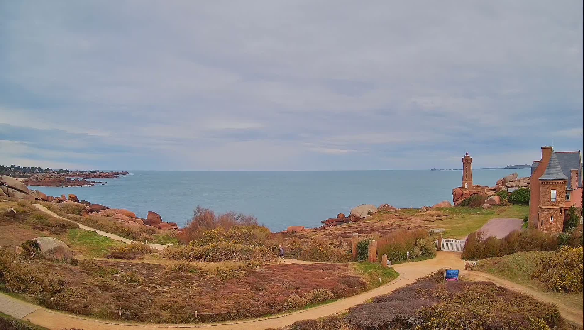 A coastal path winds through low-lying vegetation toward a lighthouse and a stone building overlooking a calm sea under a cloudy sky.