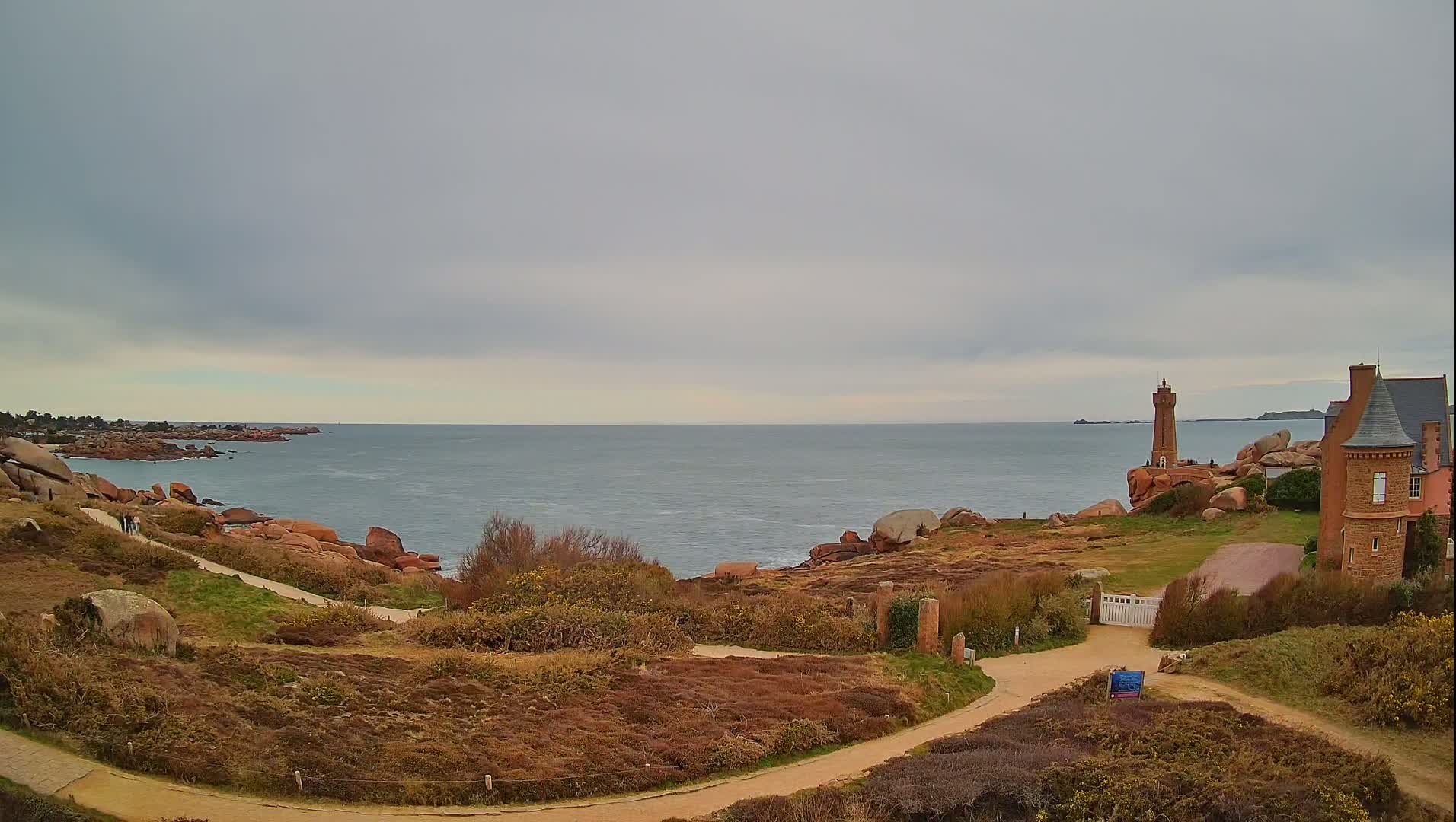 A coastal scene under a cloudy sky shows a lighthouse and a stone building overlooking a rocky coastline and calm ocean.