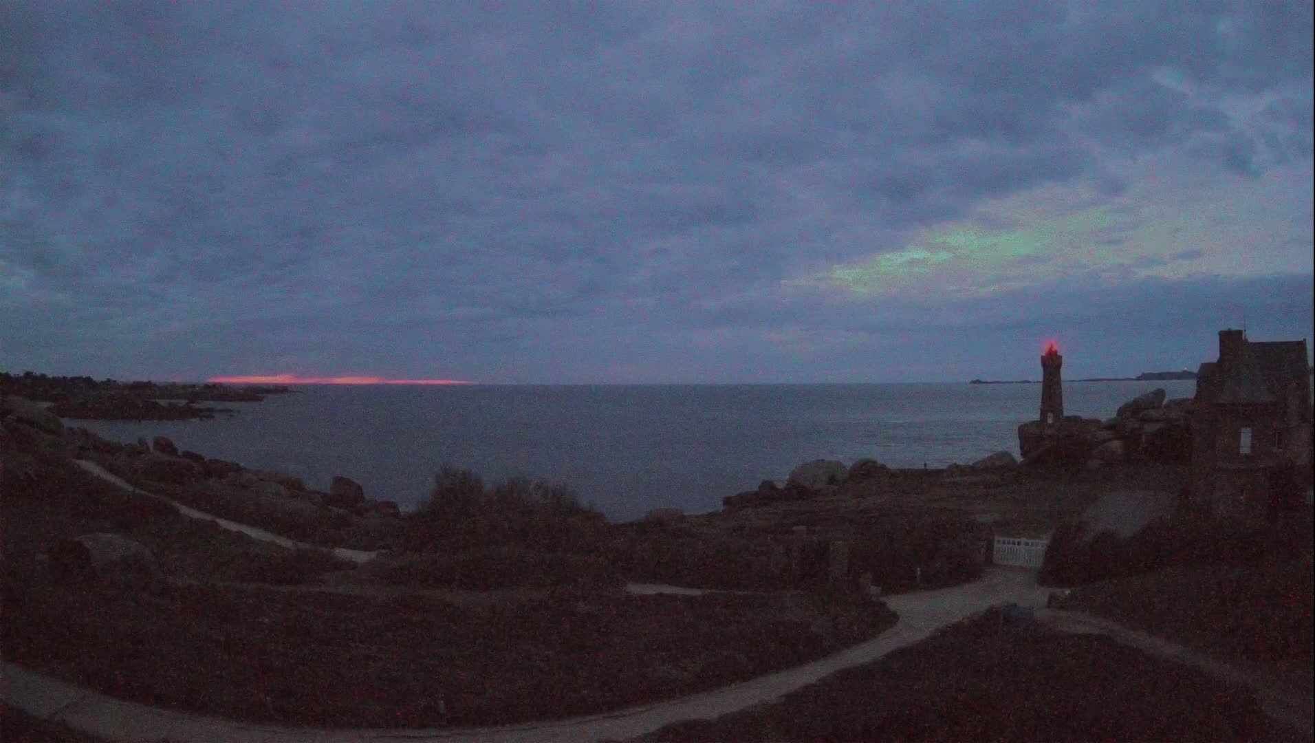 A rocky coastline at twilight with a lit lighthouse and a building near the water under a cloudy sky showing a faint pinkish glow on the horizon.