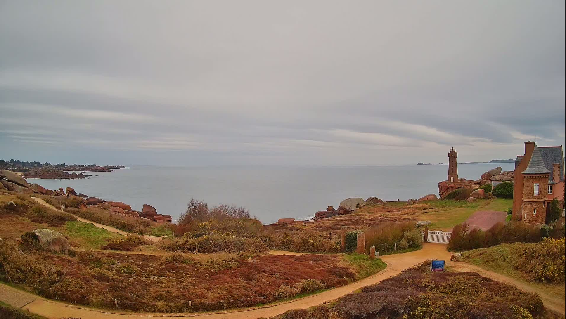 A coastal path winds through low-lying vegetation toward a lighthouse and stone house overlooking a calm, gray sea under a cloudy sky.