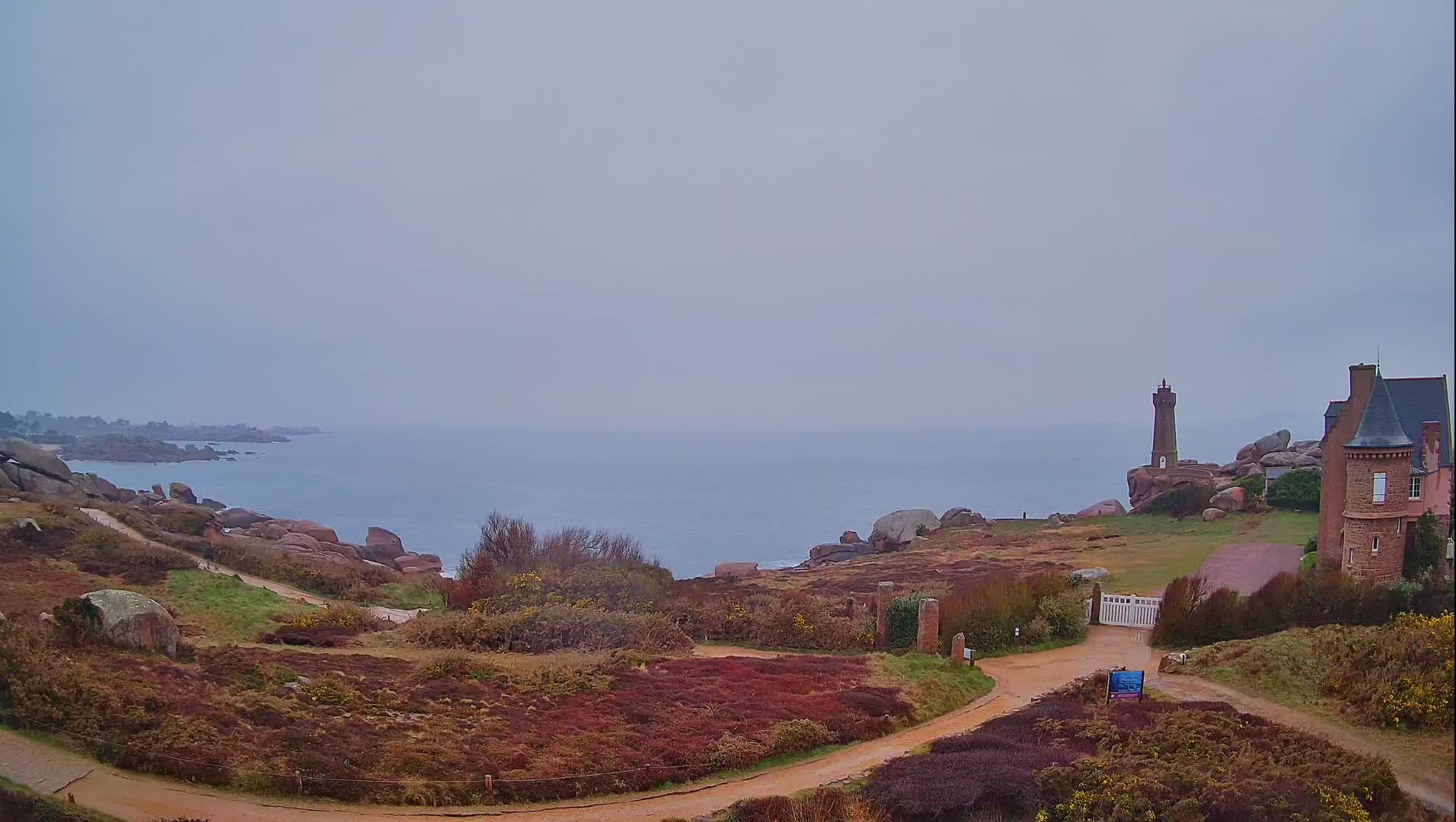 A coastal path winds through low-lying vegetation toward a stone house and lighthouse overlooking a calm, gray sea under an overcast sky.
