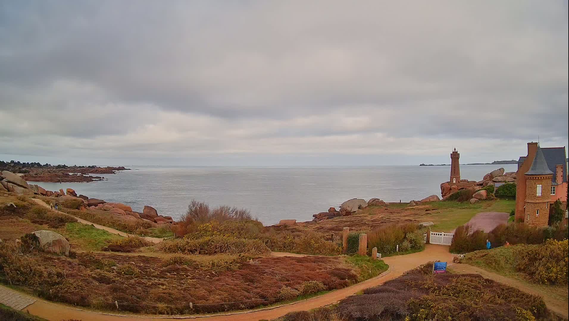 A coastal scene under a cloudy sky shows a brick lighthouse and house overlooking a calm sea and rocky shoreline with a dirt path winding through low-lying vegetation.