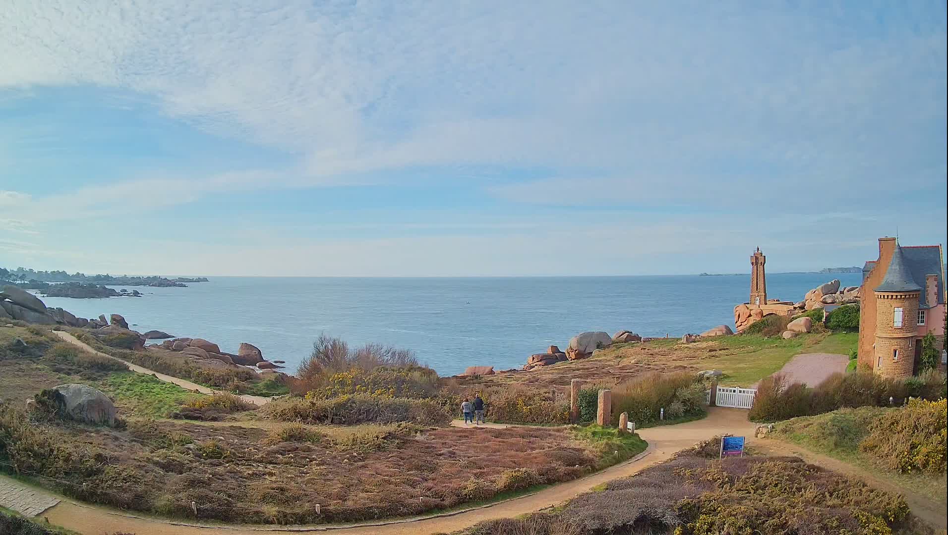 A coastal path winds through low-lying vegetation toward a stone house and a small lighthouse overlooking a calm ocean under a mostly clear sky.