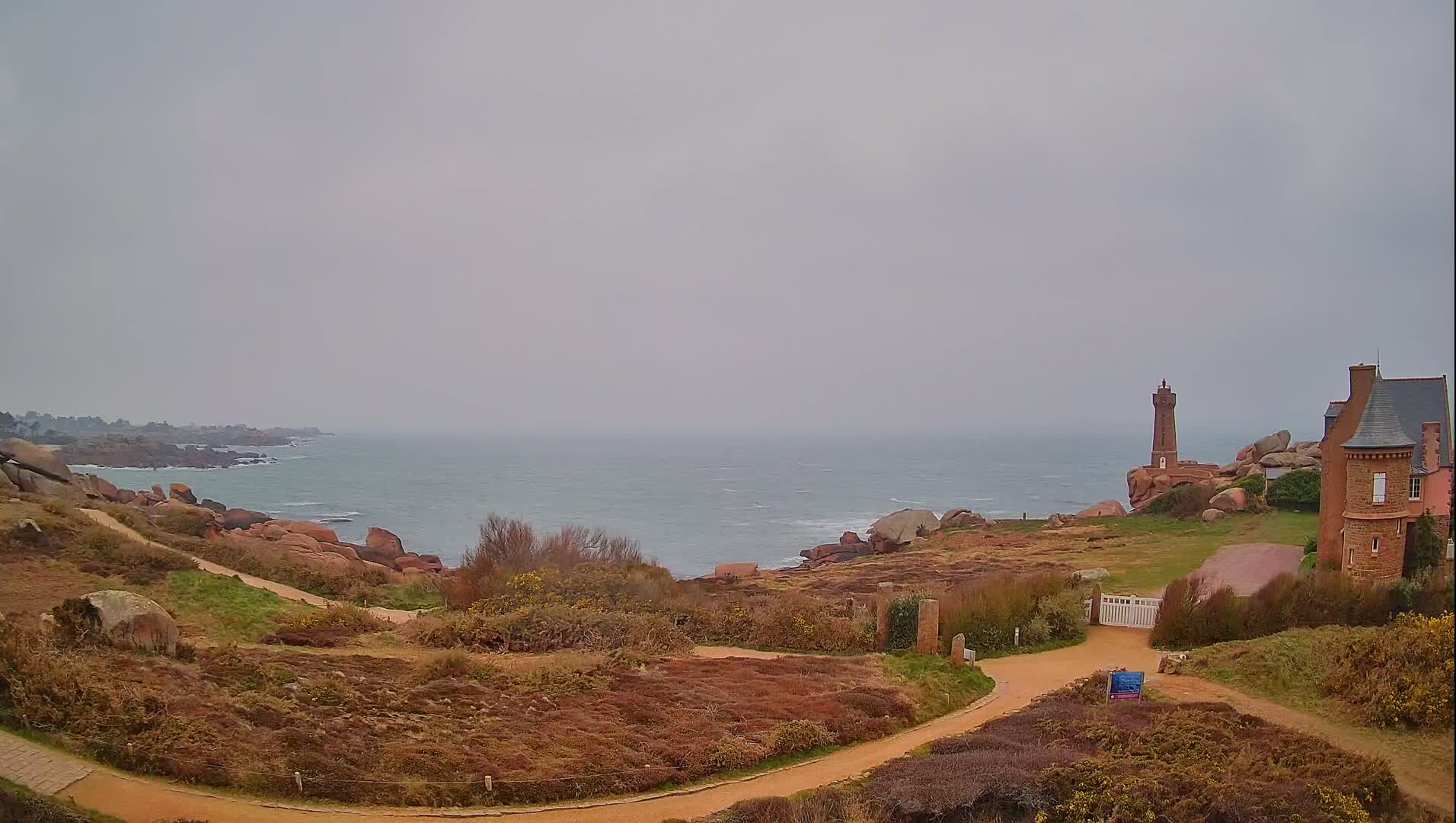 A winding path leads to a lighthouse and a stone house overlooking a rocky, overcast coastline.