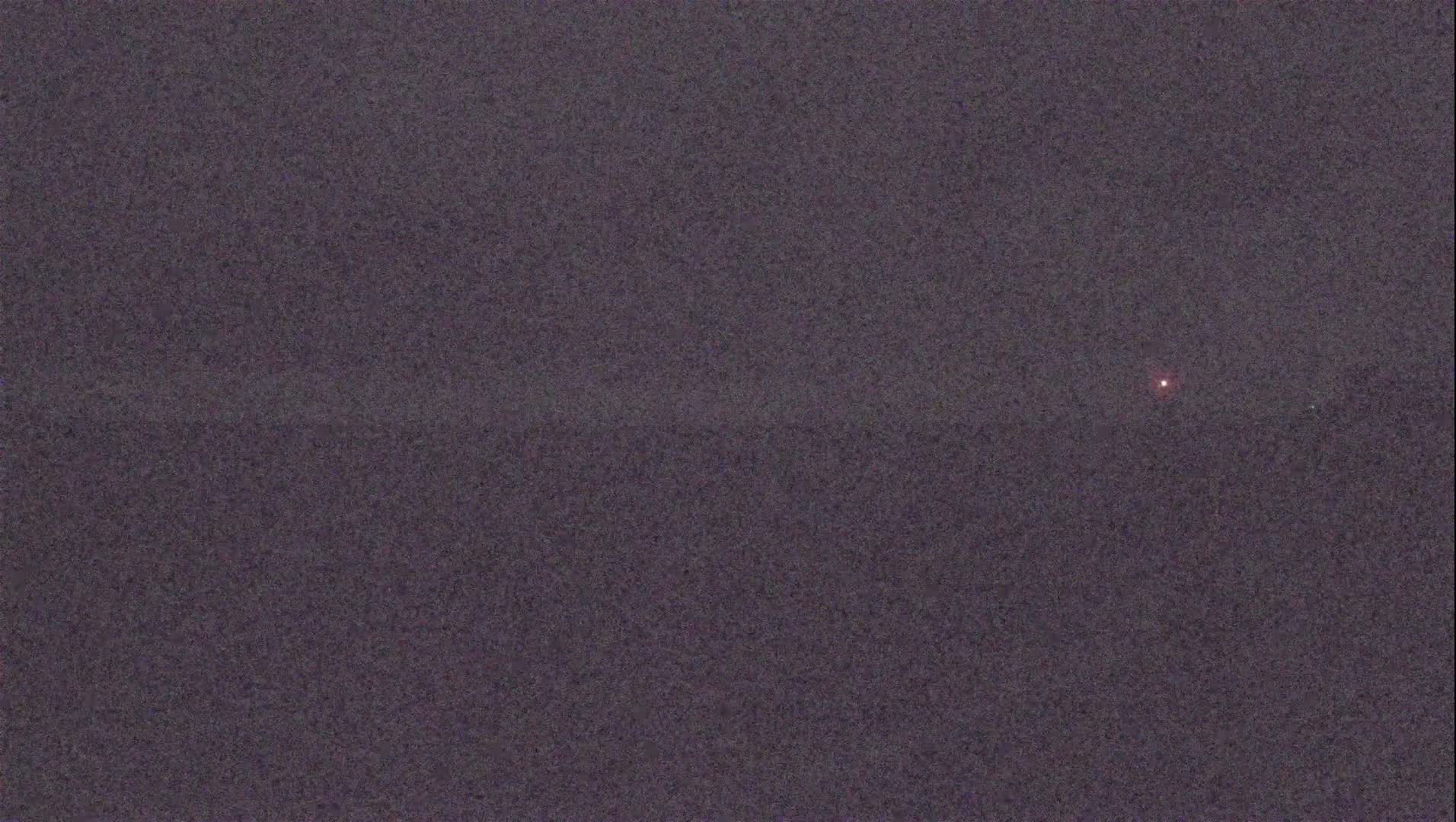 A dimly lit, dark gray sky at night shows a single, small, reddish light in the distance.