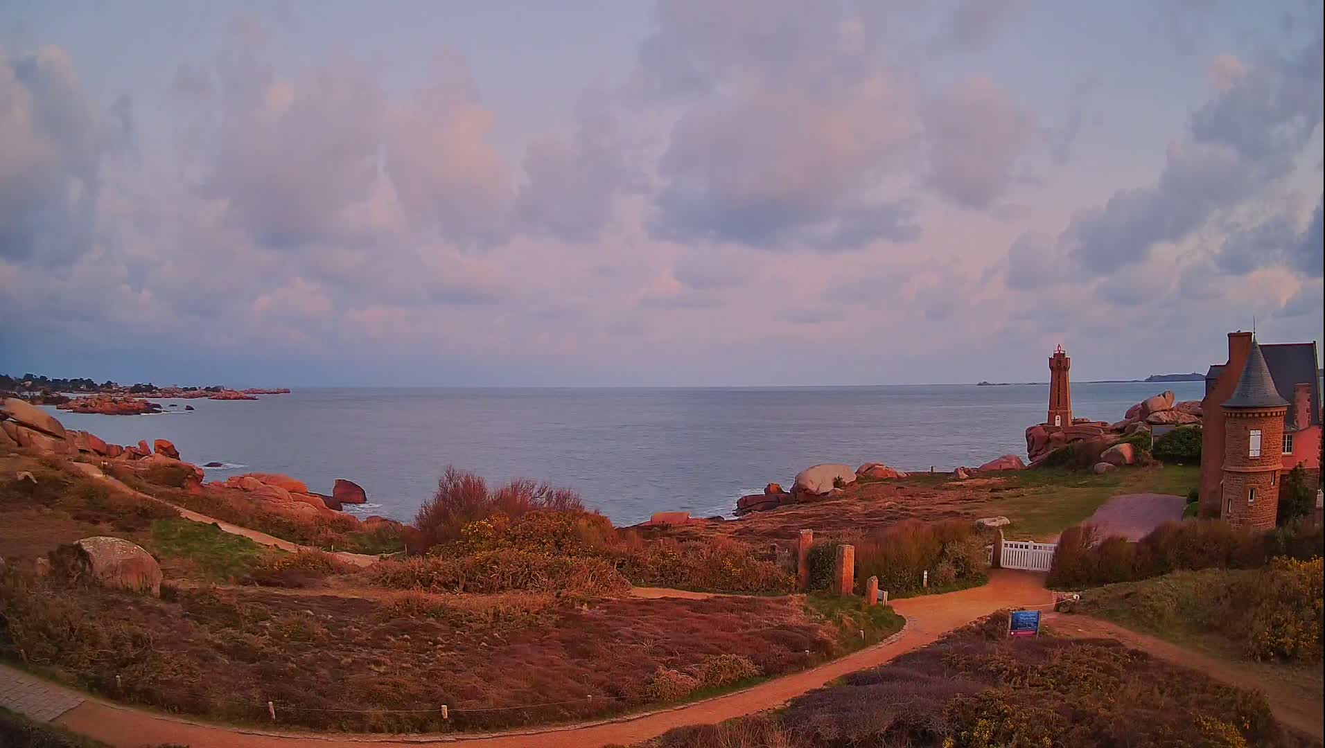 A coastal path winds through pinkish-purple heather towards a lighthouse and a stone house overlooking a calm sea under a partly cloudy, pale sky.