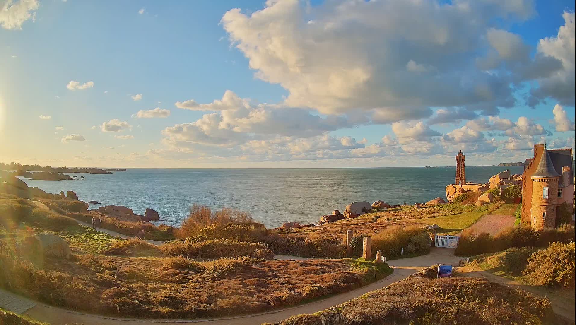 A partly cloudy sunset illuminates a coastal scene with a stone building, a lighthouse, and a winding path along a rocky shoreline.