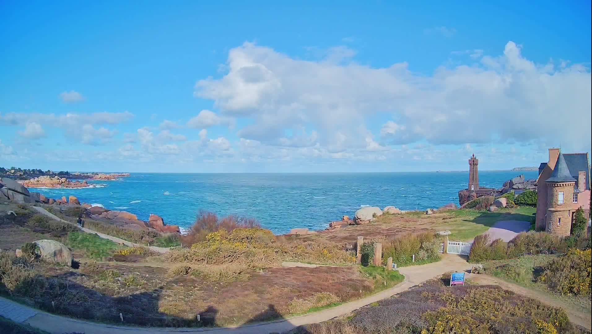 A partly cloudy day shows a rocky coastline with a lighthouse and a stone building overlooking a blue ocean with small waves.