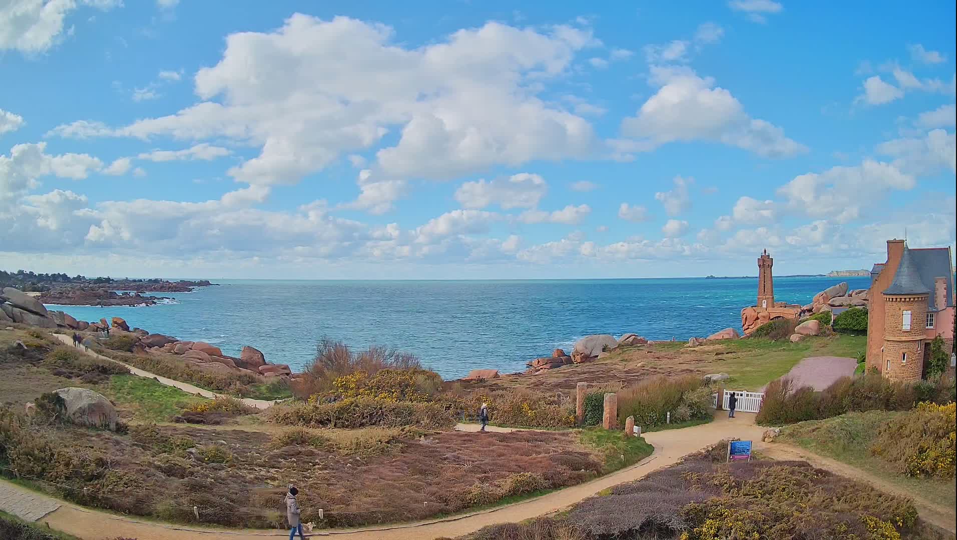 A coastal path winds through low-lying vegetation toward a lighthouse and a stone building overlooking a blue ocean under a partly cloudy sky.