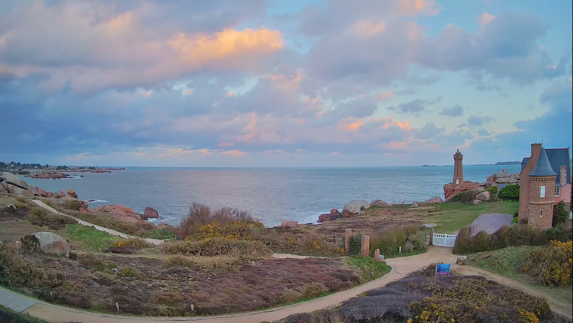 A coastal path winds through low-lying vegetation toward a stone house and a small lighthouse overlooking a calm sea under a partly cloudy sky with soft, pastel hues.
