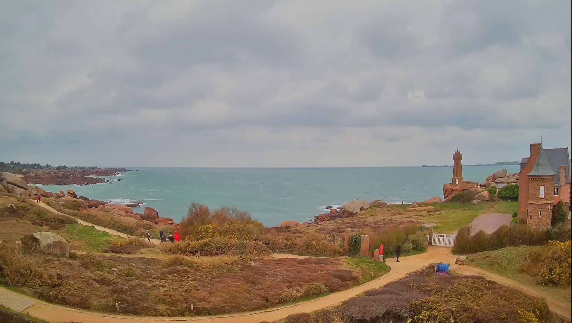 A coastal scene under a cloudy sky shows a rocky shoreline, a lighthouse, a stone building, and several people walking along paths.