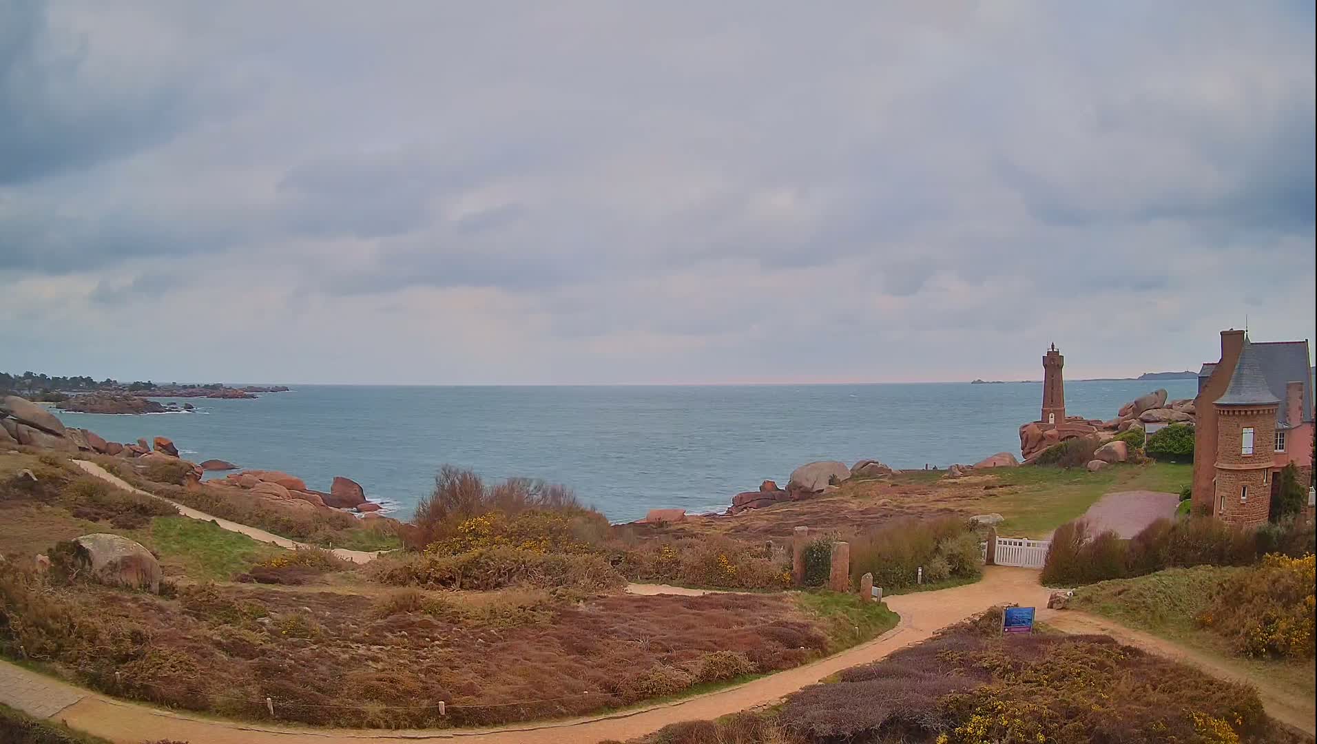 A winding path leads to a gate near a stone building overlooking a calm sea with a lighthouse on a rocky point under an overcast sky.