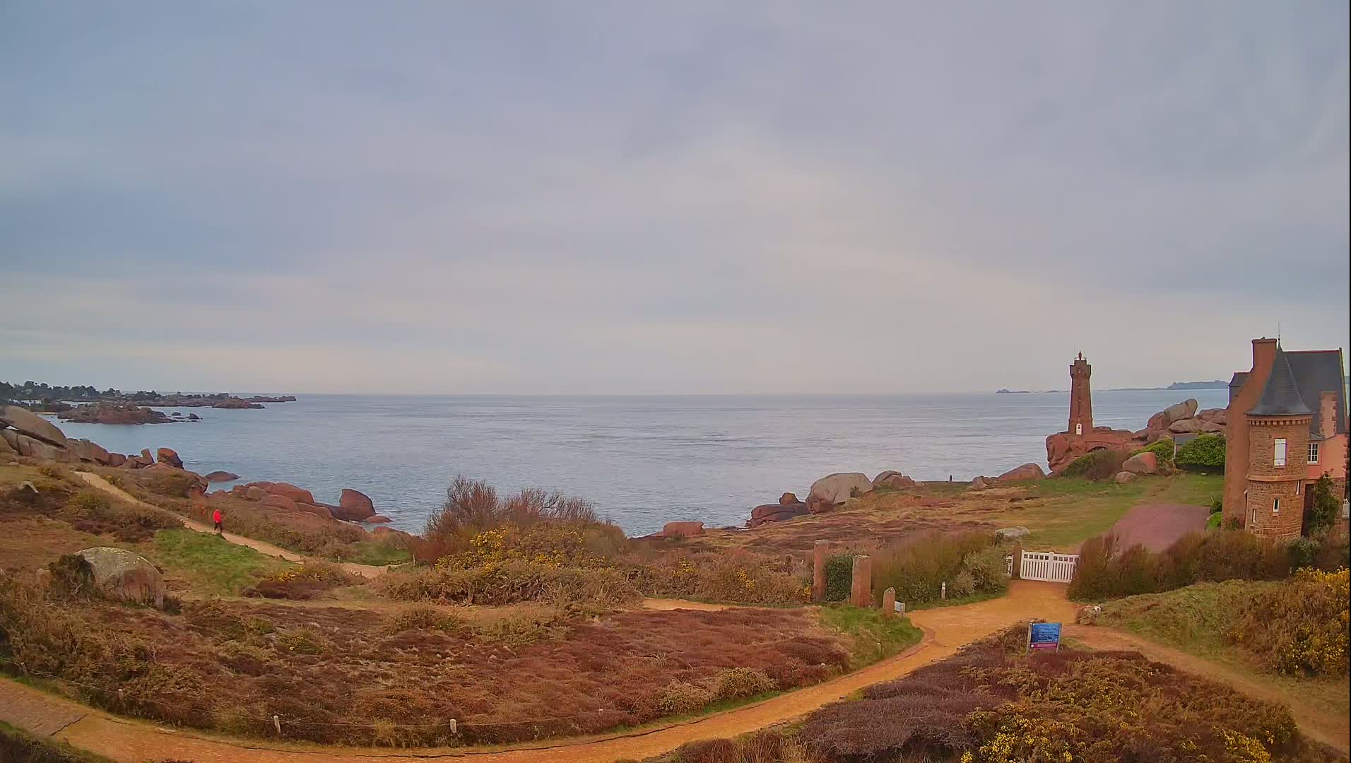 A coastal scene under an overcast sky shows a person walking a path near a brick lighthouse and house overlooking a calm sea with rocky shores.