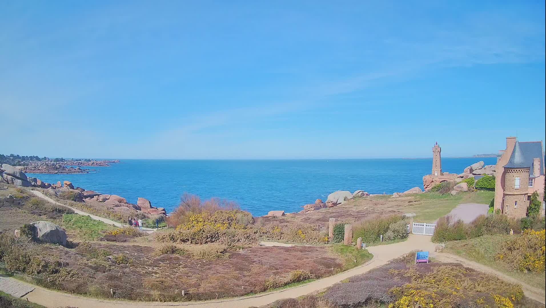 A coastal scene on a sunny day shows a lighthouse and a stone building overlooking a calm blue ocean, with pinkish-red rocks and low-lying vegetation in the foreground.