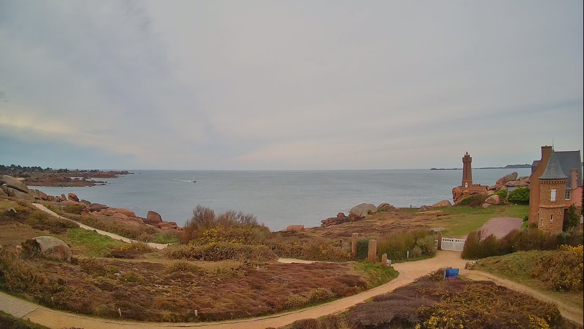 A coastal scene on an overcast day shows a rocky shoreline with a lighthouse and a stone building, winding paths, and low vegetation leading to a calm sea with a small boat in the distance.