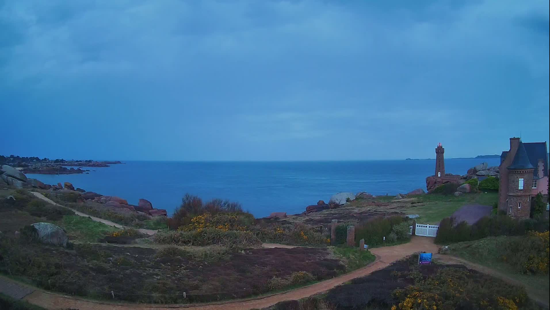 A coastal scene under a mostly clear, twilight sky shows a lighthouse and a stone house on a rocky outcropping overlooking a calm sea.