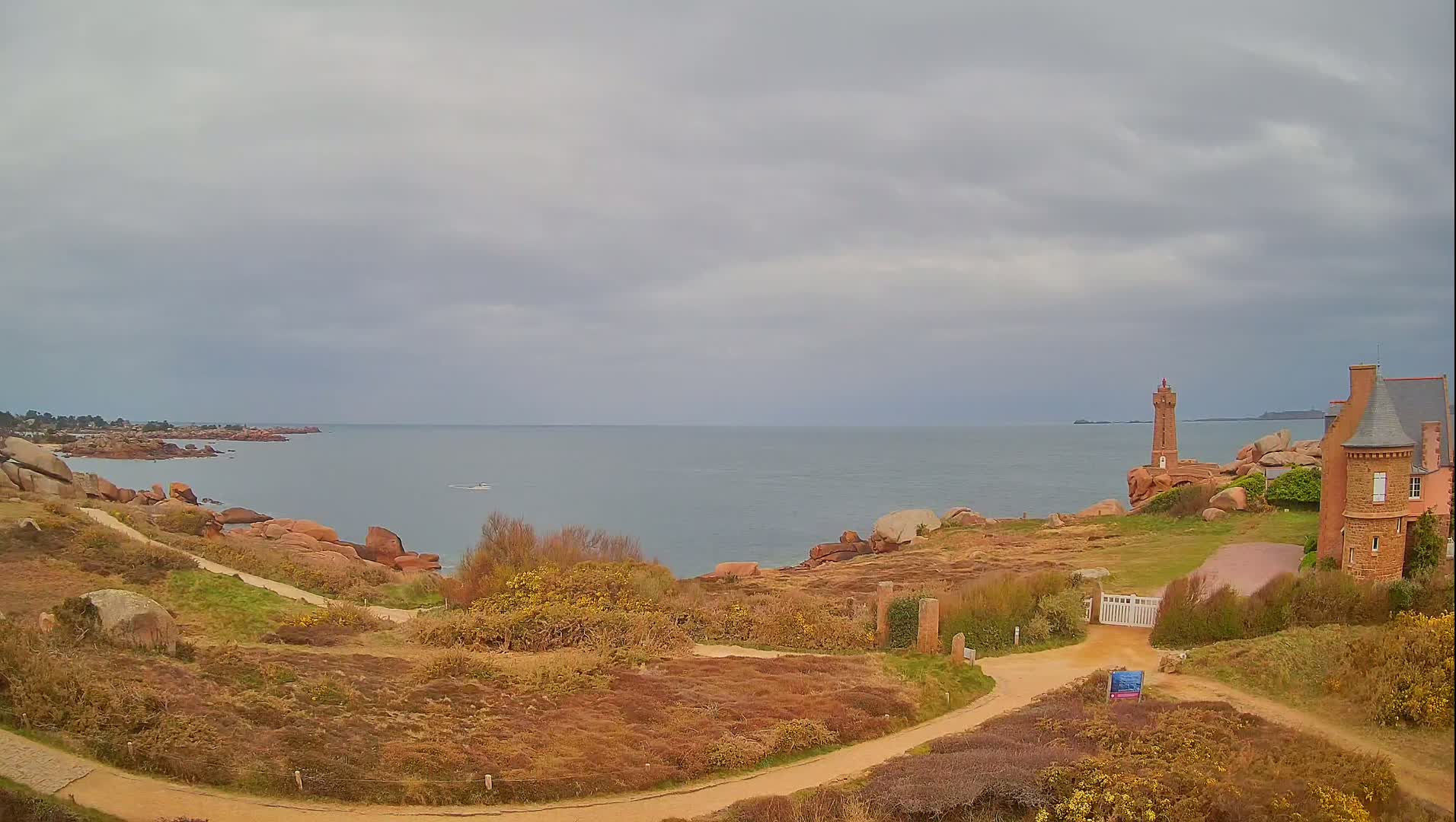 A coastal scene under an overcast sky shows a dirt path winding toward a gated entrance to a stone house near a rocky shoreline with a small lighthouse visible in the distance.