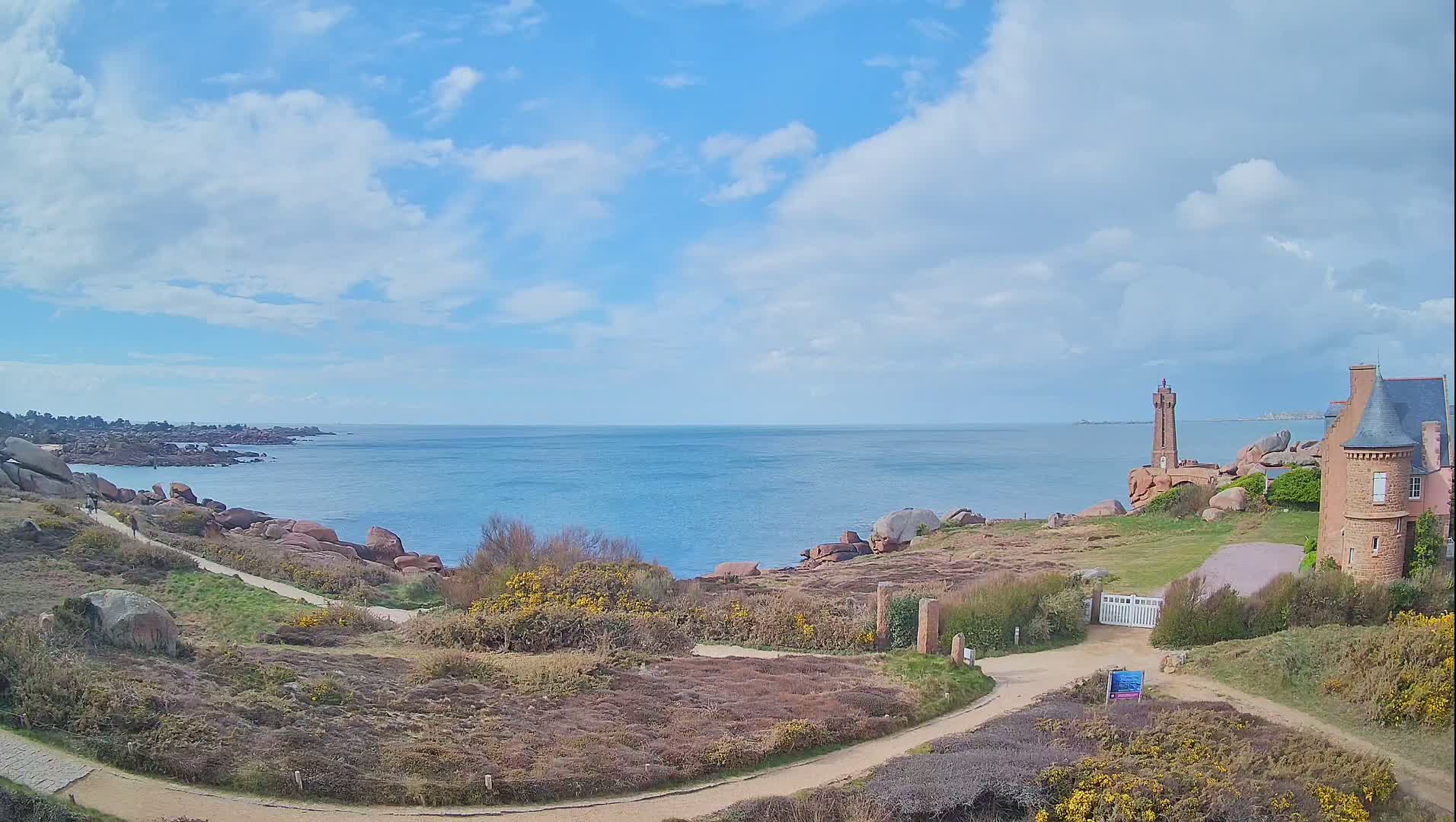 A coastal scene under a partly cloudy sky shows a rocky shoreline with a lighthouse and a stone building, connected by a winding path.