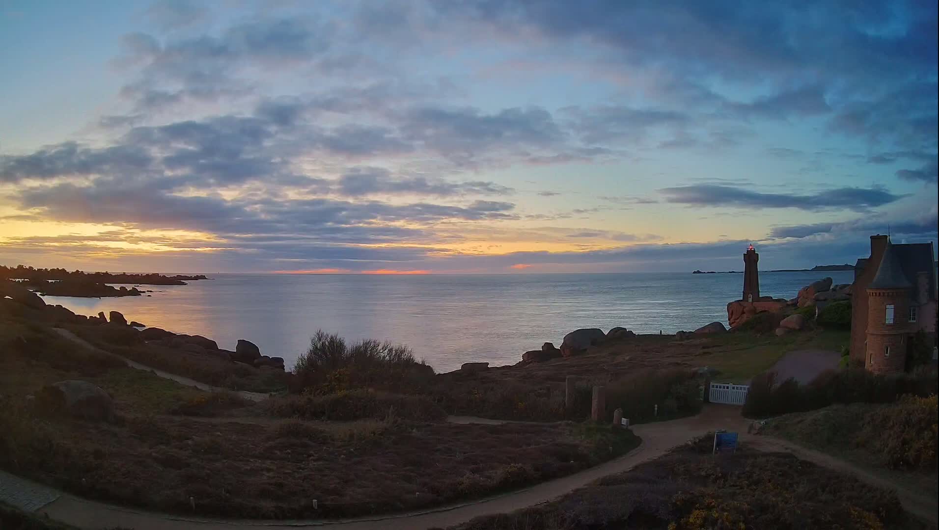 A coastal scene at sunset shows a lighthouse and a stone building on a rocky headland overlooking a calm sea under a partly cloudy sky.