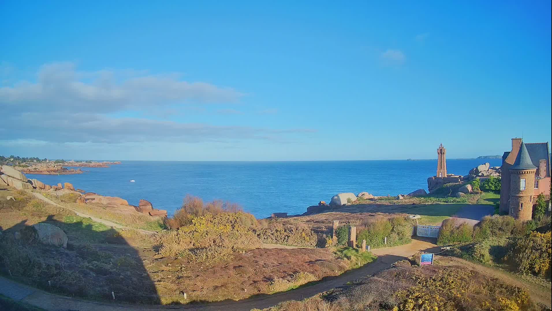 A coastal scene on a clear, sunny day shows a rocky shoreline, a small lighthouse, a stone house, and a calm blue ocean extending to the horizon.