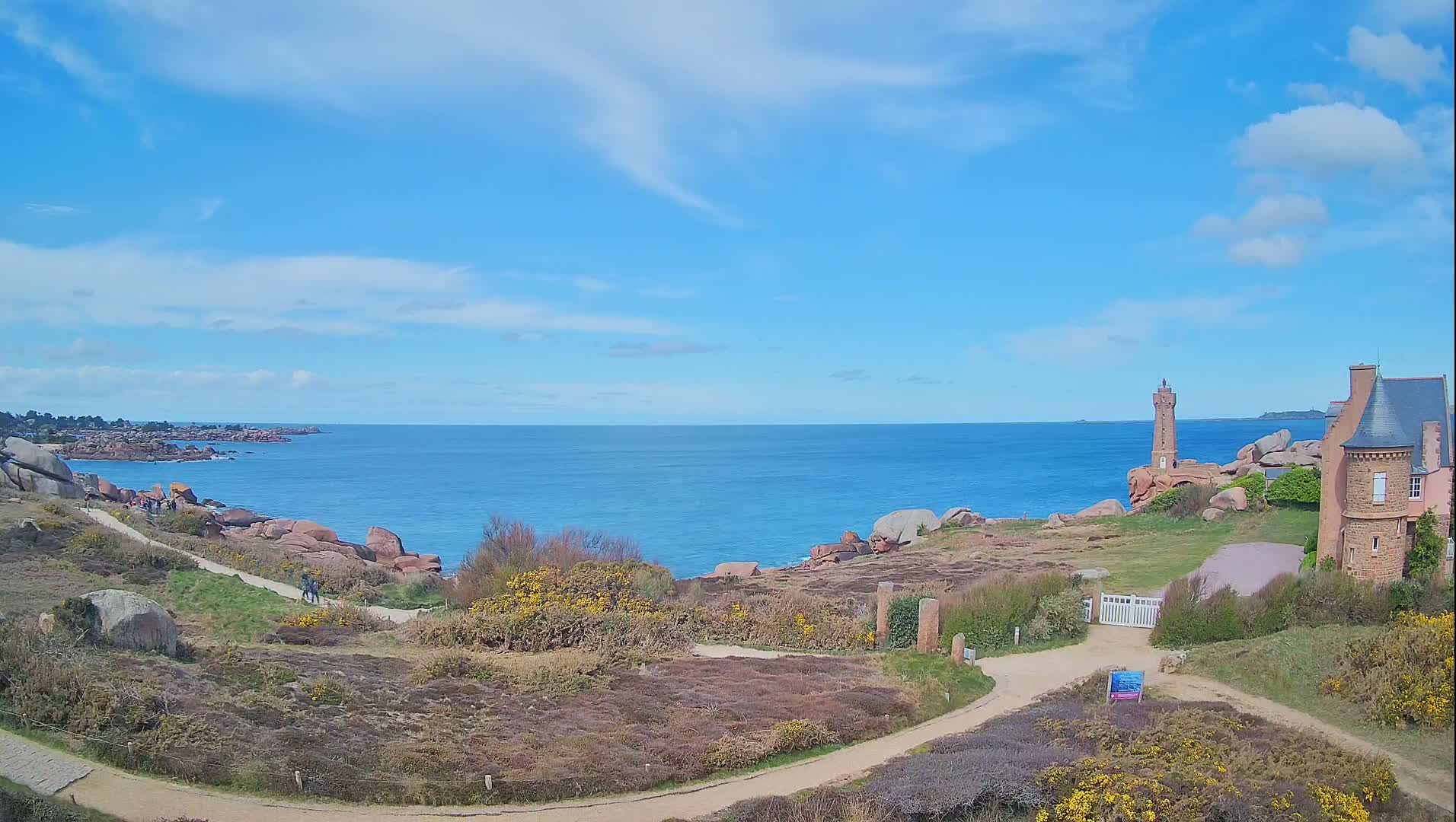 A coastal path winds through low-lying vegetation toward a lighthouse and a stone house overlooking a calm blue ocean under a mostly sunny sky.