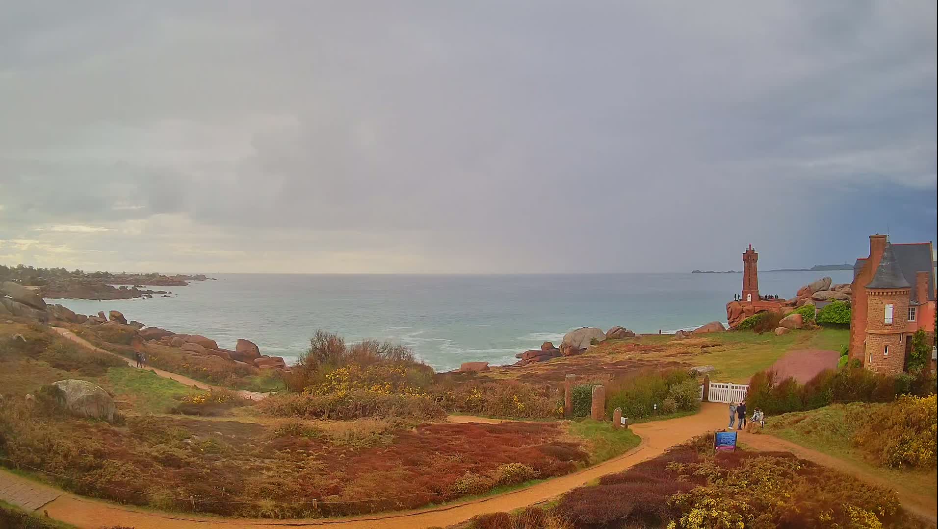 A brick lighthouse stands on a rocky outcrop overlooking a calm, gray-toned ocean under a cloudy sky, with a path leading to a nearby stone building.