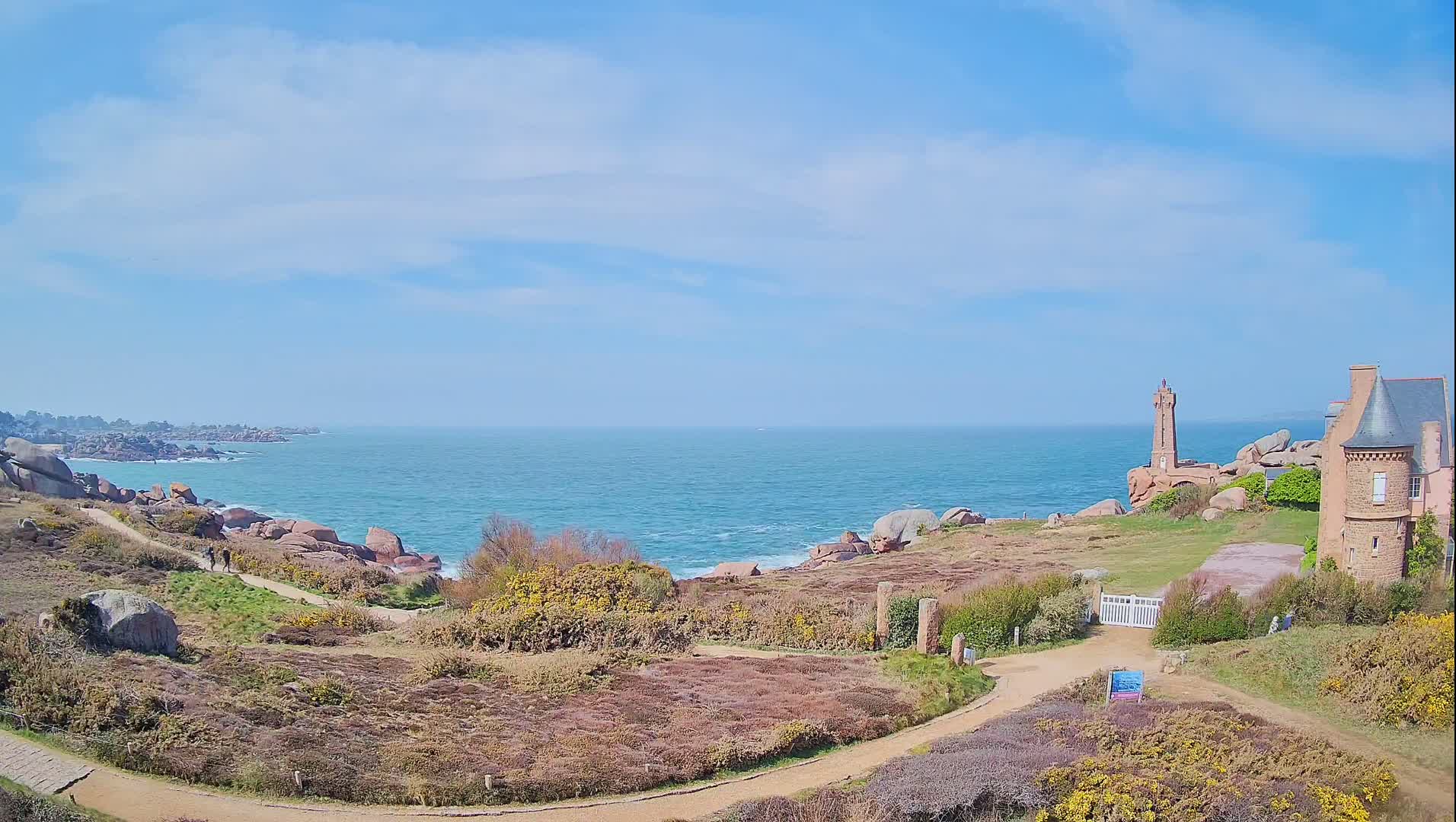 A coastal scene under a mostly sunny sky shows a stone building near a lighthouse overlooking a rocky shoreline and blue ocean.