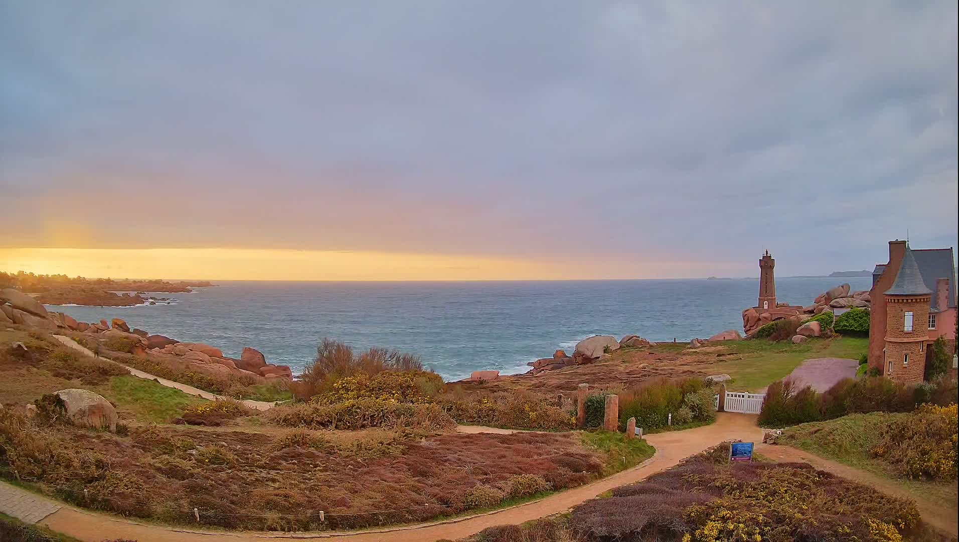 A pink and orange sunset over a rocky coastline with a lighthouse and a stone house, under a partly cloudy sky.
